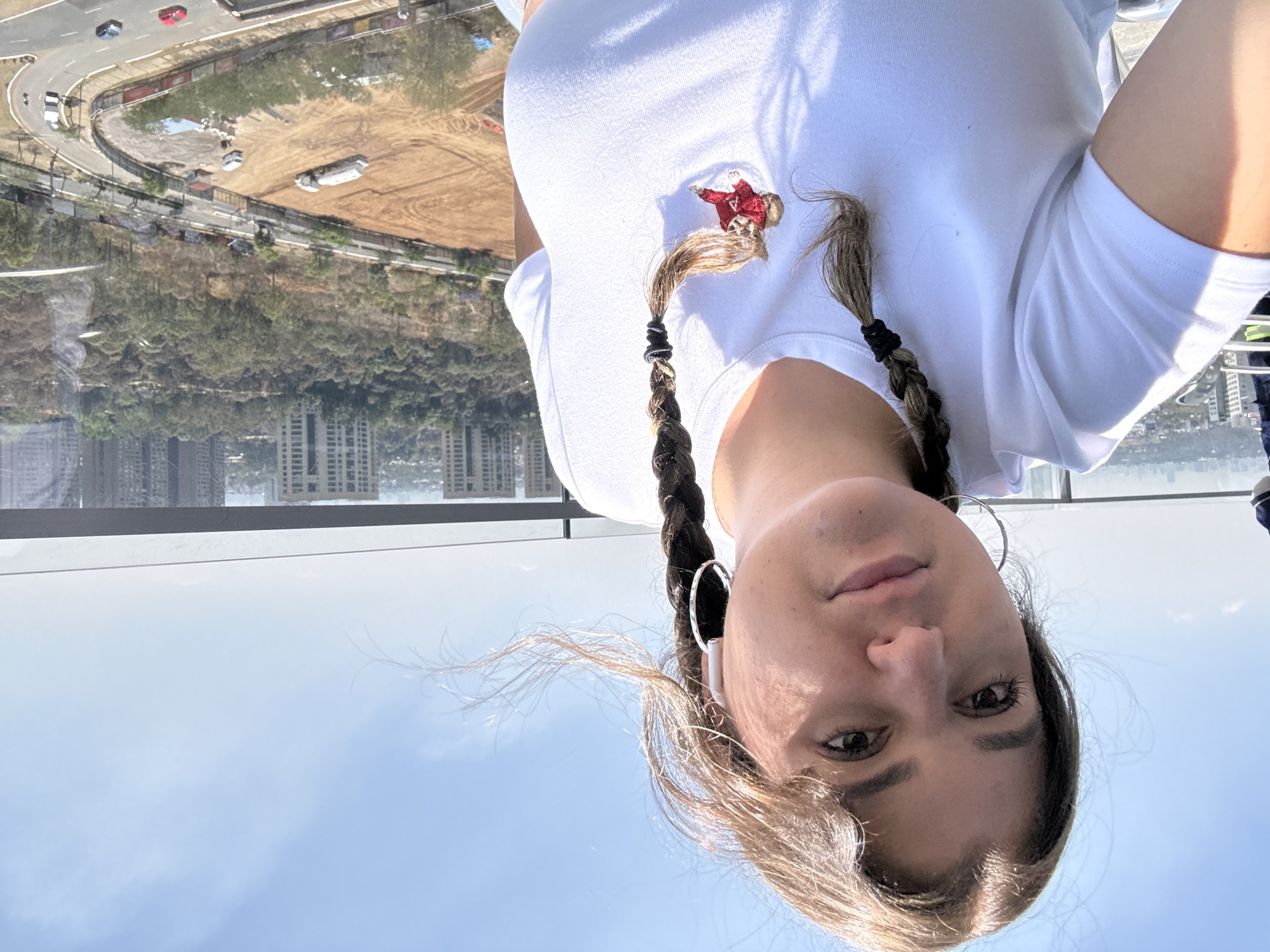 A woman is looking directly at the camera. She has light skin and brown eyes. Her blonde hair is styled in two braids with black elastics. She is wearing silver hoop earrings and a white t-shirt with a small embroidered figure of a person in a red coat.
The background shows a panoramic view through a glass partition. Below is a view of Mexico City, Mexico. There is a road with moving vehicles, bare land with construction, trees, and buildings. The sky is visible and clear in some areas.