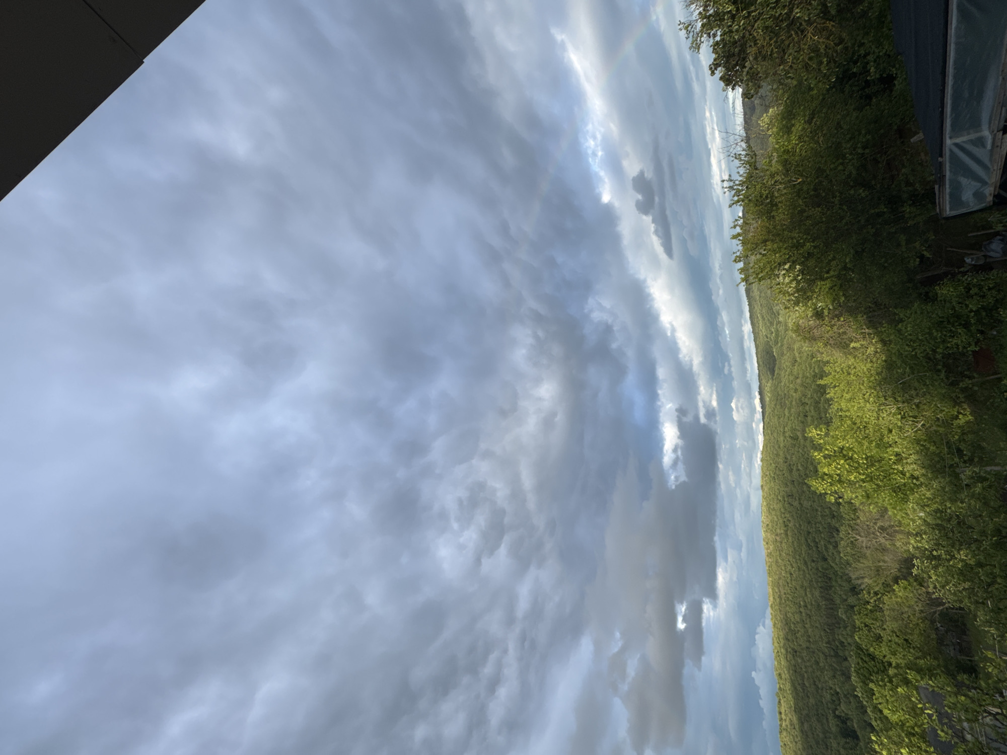 The image shows a landscape view oriented vertically. The sky takes up the majority of the frame, filled with dense, gray clouds, with hints of sunlight filtering through. A faint rainbow is visible near the horizon. On the right side, lush green trees and vegetation cover a sloping hillside. The edge of a structure is visible at the top left. A building and windows can be seen on the top right of the image. The location is likely in Konstantinovo, Bulgaria, based on the request. No people are visible.