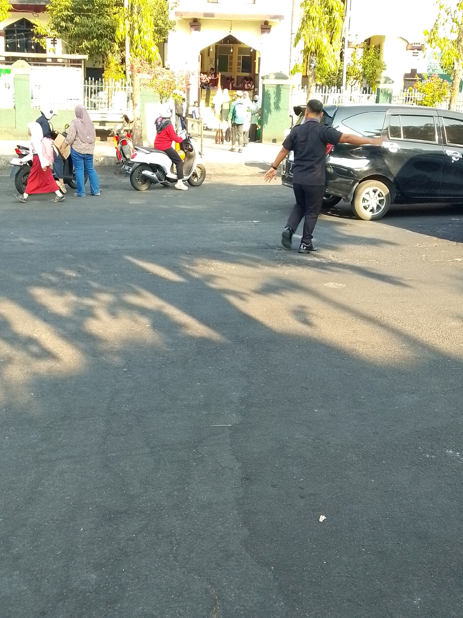 In Jember, Indonesia, at the entrance of a building that appears to be a mosque or school, several individuals are gathered. A security guard in a black uniform is directing traffic near a black car. In the foreground, two young girls in red uniforms and headscarves are walking, one holding a pink backpack. Another woman in a light brown hijab and blue jeans is standing next to a motorbike. To the right, a person in a red shirt and black pants is seated on a white motorbike. Trees with yellowing leaves are visible in the background, and shadows from the trees are cast across the asphalt road.
