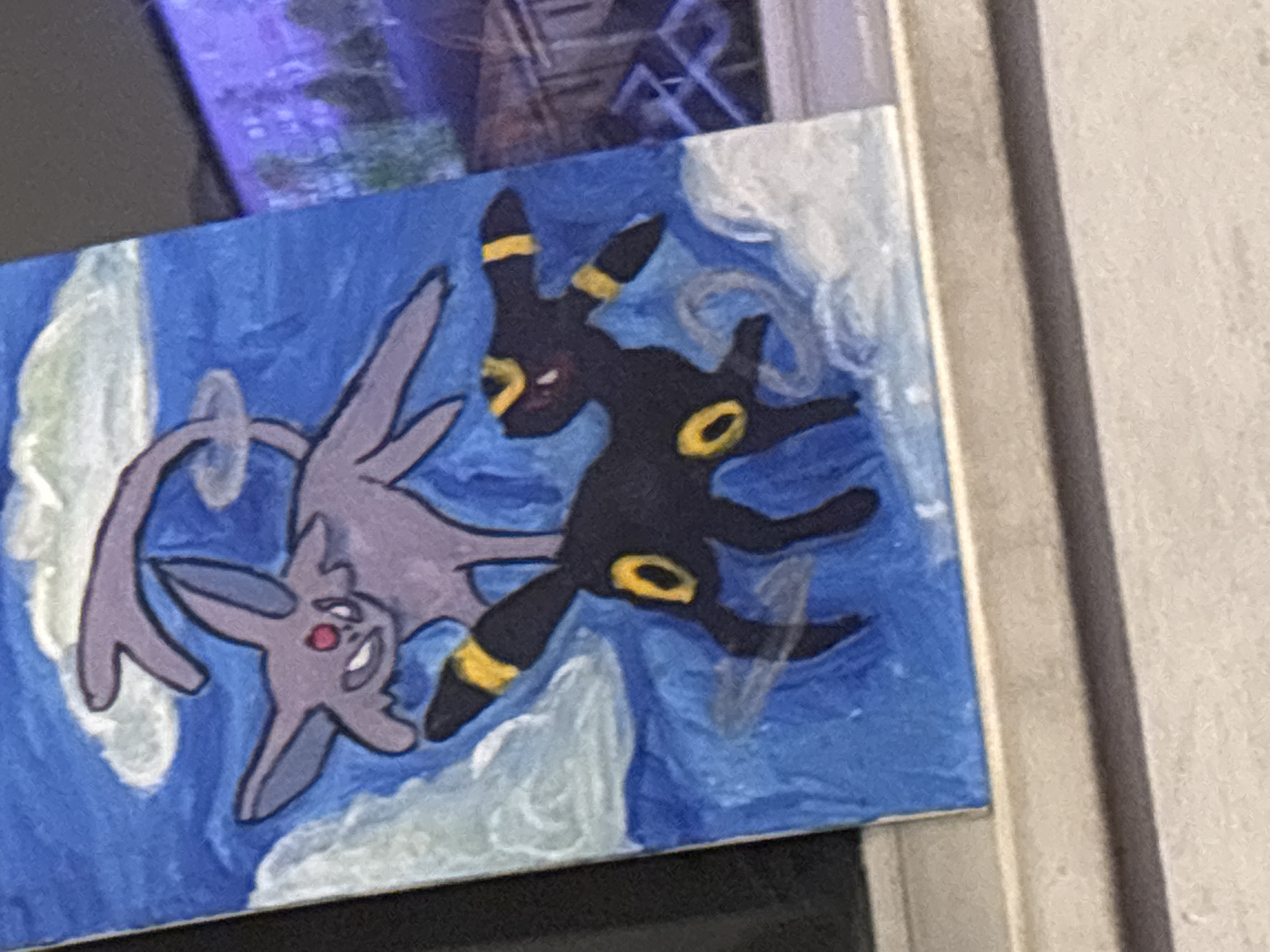 A painting depicting the Pokémon Espeon and Umbreon is visible in the image. The painting is on a blue background with white swirls. Espeon, a purple Pokémon, is on the left, and Umbreon, a dark Pokémon with yellow rings, is on the right. The painting appears to be on a canvas or board. The environment appears to be indoors, possibly near a window or door. The city and country of Buenos Aires, Argentina are not visually discernible from the image.
