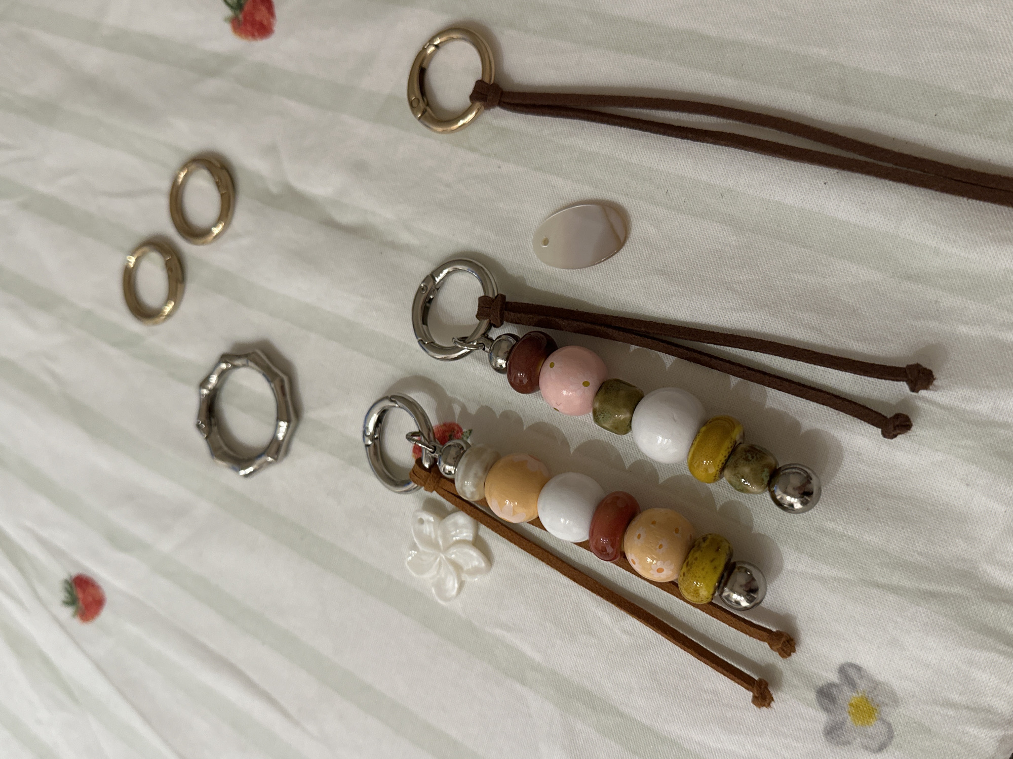 The image shows an array of craft materials arranged on a white surface with a faint striped pattern. There are two gold-colored hoop earrings, a silver-colored octagonal ring, a golden ring attached to two brown leather cords, two keychains with silver-colored clasps and beaded designs attached to brown leather cords, and a small white shell bead. One of the keychains also has a small white flower charm attached. The image was not taken in Šušanj, Montenegro, and no people, subjects, objects, actions, interactions, or environment are relevant to that location.