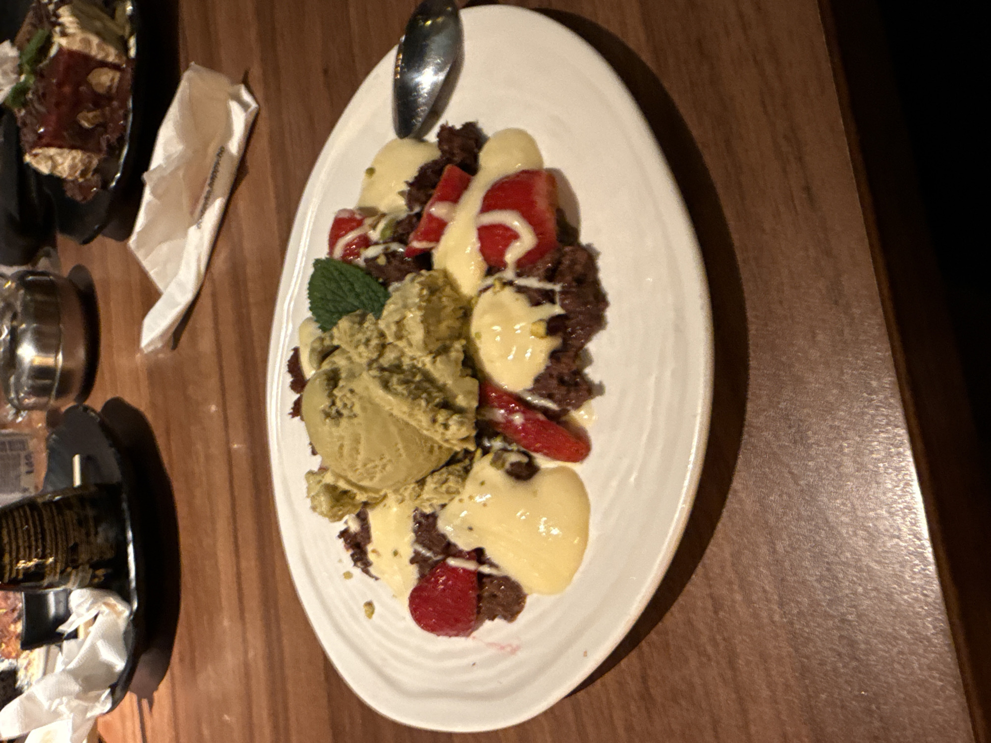 The image shows a dessert served on a white plate, likely taken in a restaurant or similar establishment. The dessert consists of what appears to be a chocolate crumble base, topped with scoops of pistachio ice cream, fresh strawberries and raspberries, and a white sauce, possibly a custard or vanilla sauce. A sprig of mint garnishes the dessert. A spoon rests on the plate. The dessert sits on a wooden table. Other plates with food are partially visible in the background, one containing a layered dessert. A folded white napkin also appears next to one of the plates in the background. There is no indication that the image was taken in Burgas, Bulgaria. There are no people visible in the image.
