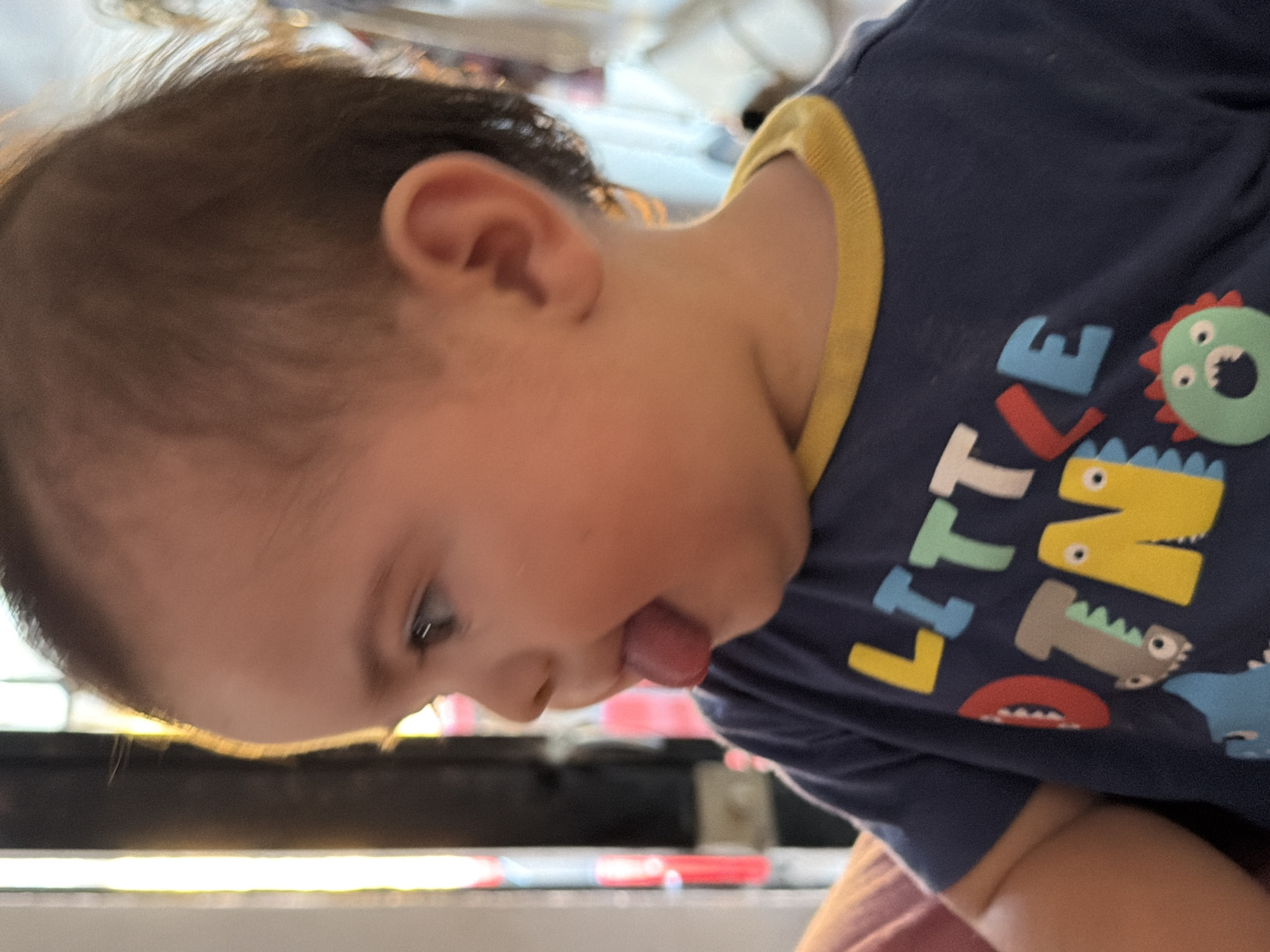 A young child is pictured close-up, sticking out their tongue. The child is wearing a dark blue t-shirt with a yellow collar and colorful dinosaur-themed lettering that reads "LITTLE DINO." The child's hair is brown and the skin is light. The background is out of focus. There is no verifiable information to place this image in Reforma, Mexico.