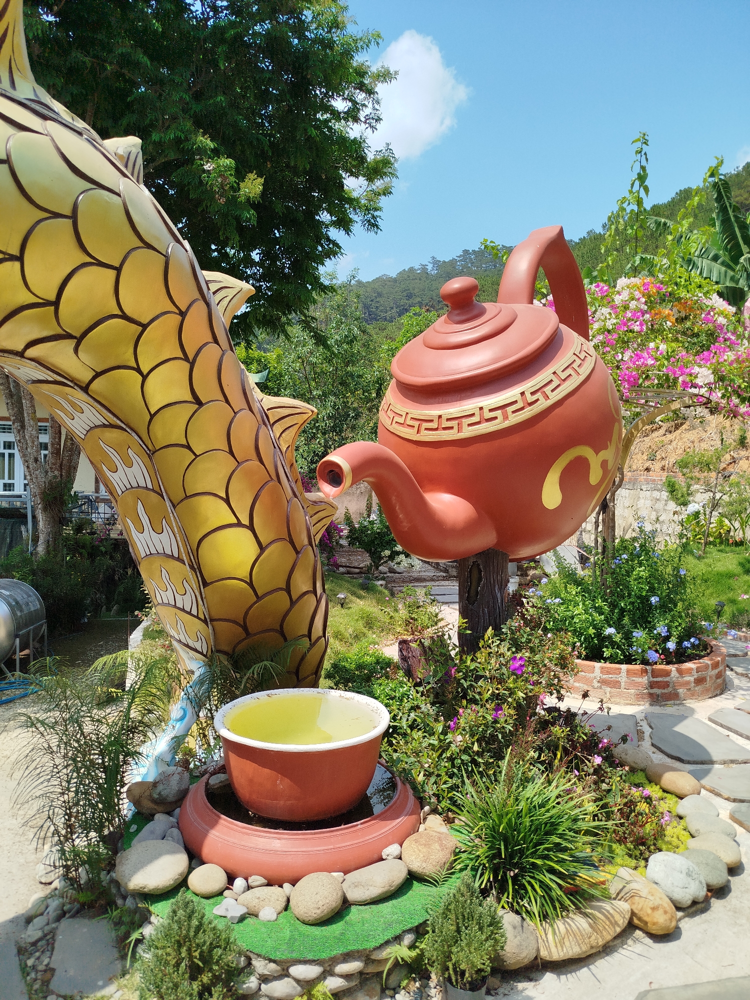 The scene depicts an outdoor garden display in Diom A, Vietnam. A large, golden dragon sculpture is positioned next to an oversized, rust-colored teapot sculpture. The teapot appears to be pouring into a bowl placed below, presumably creating a fountain feature. The garden is decorated with various plants, flowers, and rocks, creating a whimsical and serene atmosphere. The sky is blue with scattered clouds.