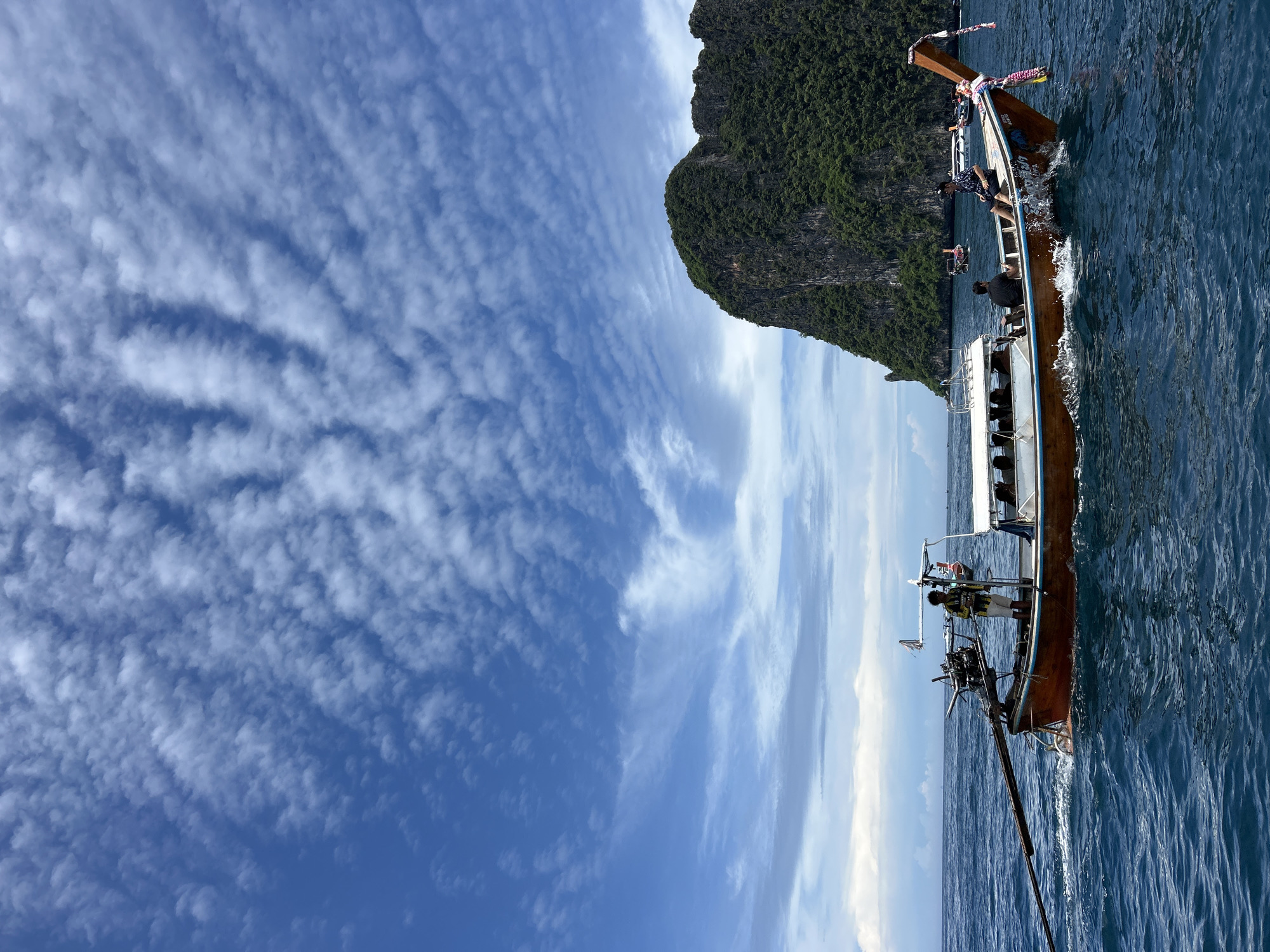 A longtail boat navigates the deep blue waters of Ao Nang, Thailand, under a vibrant blue sky dotted with textured cumulus clouds. The wooden boat, painted orange-brown with a light blue stripe and a decorative colorful garland on its bow, moves from left to right, creating white splashes around its hull. Five individuals are visible on board: one person, dressed in a yellow and black striped shirt, stands at the stern near the long-shaft engine; two passengers are seated under a white canopy in the mid-section; and two other individuals are seated towards the bow, one wearing a black shirt and the other a patterned dark shirt. In the background to the right, a large, dark green limestone karst island, heavily vegetated, rises steeply from the sea. Several other longtail boats are visible in the distance, scattered across the water near the island and further out. The horizon line on the left shows a distinct separation between the ocean and the sky, where lighter, wispy clouds are present closer to the water.