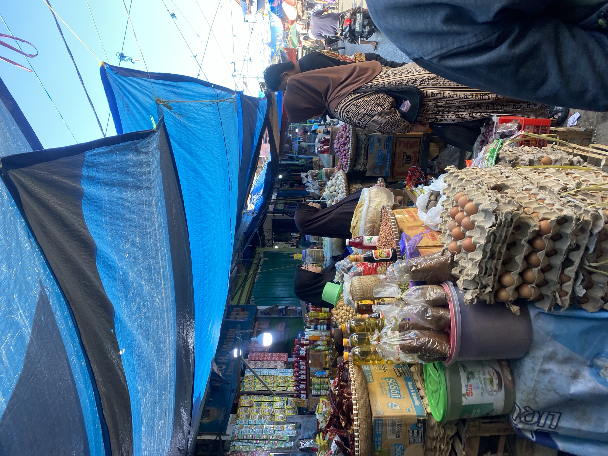 This image captures a bustling market scene in Desa Tunggul Irang, Indonesia. A vibrant blue tarp, supported by dark fabric, stretches overhead, creating a shaded area for vendors and shoppers. The market stalls are laden with a variety of goods, including dried chilies, bottles of cooking oil and sauces, bags of nuts and grains, and stacks of eggs in cardboard cartons. Several people are visible, including two women wearing hijabs who appear to be vendors or customers, and a man in the background. One woman in a brown patterned dress is engaged in conversation with a man. The overall environment suggests a lively marketplace with abundant produce and commercial activity.