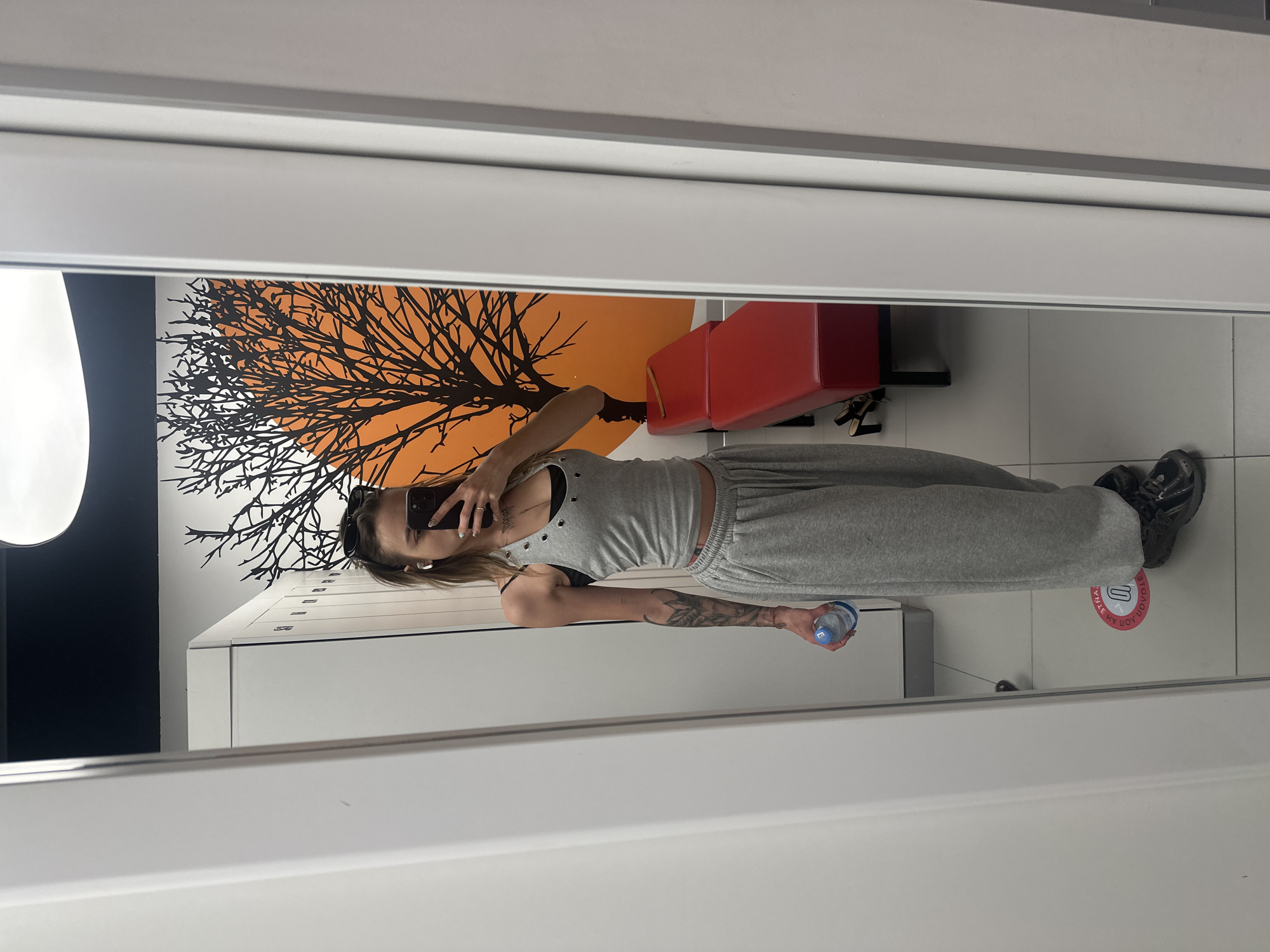 A young woman with blonde hair and sunglasses on her head is taking a mirror selfie in a locker room. She is wearing a gray tank top and matching sweatpants. She is holding a plastic water bottle in her left hand. Her right arm has a tattoo. The locker room has white lockers on the left side and a red bench on the right side. A graphic of a black tree with an orange circle behind it is on the wall. A circular red and white floor sticker is visible on the floor. The overall setting appears to be a fitness facility. It is not possible to determine the location as Moscow, Russia from the image.