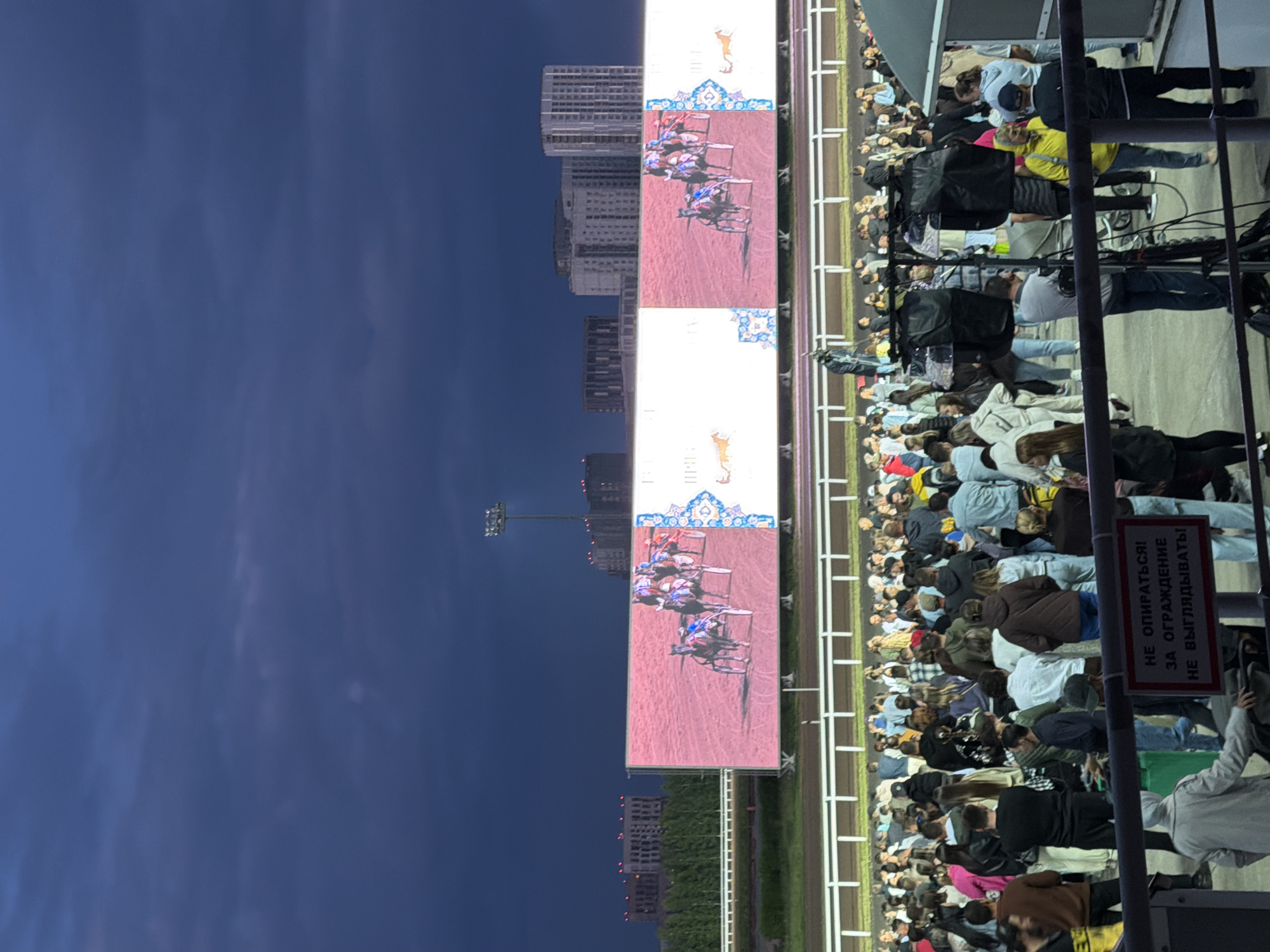 An outdoor nighttime shot shows a crowd of people watching a horse race on a large screen at a racetrack in Unknown, Unknown. The screen displays a close-up of horses and chariots racing on a reddish-brown track, with decorative elements on the sides. Above the screen, the dark, cloudy sky is illuminated by stadium lights and distant city buildings. The crowd, composed of diverse individuals, stands behind a white railing, some holding cameras and other equipment. In the foreground, a sign with Russian text is visible, advising people not to lean on the fence and not to look out.