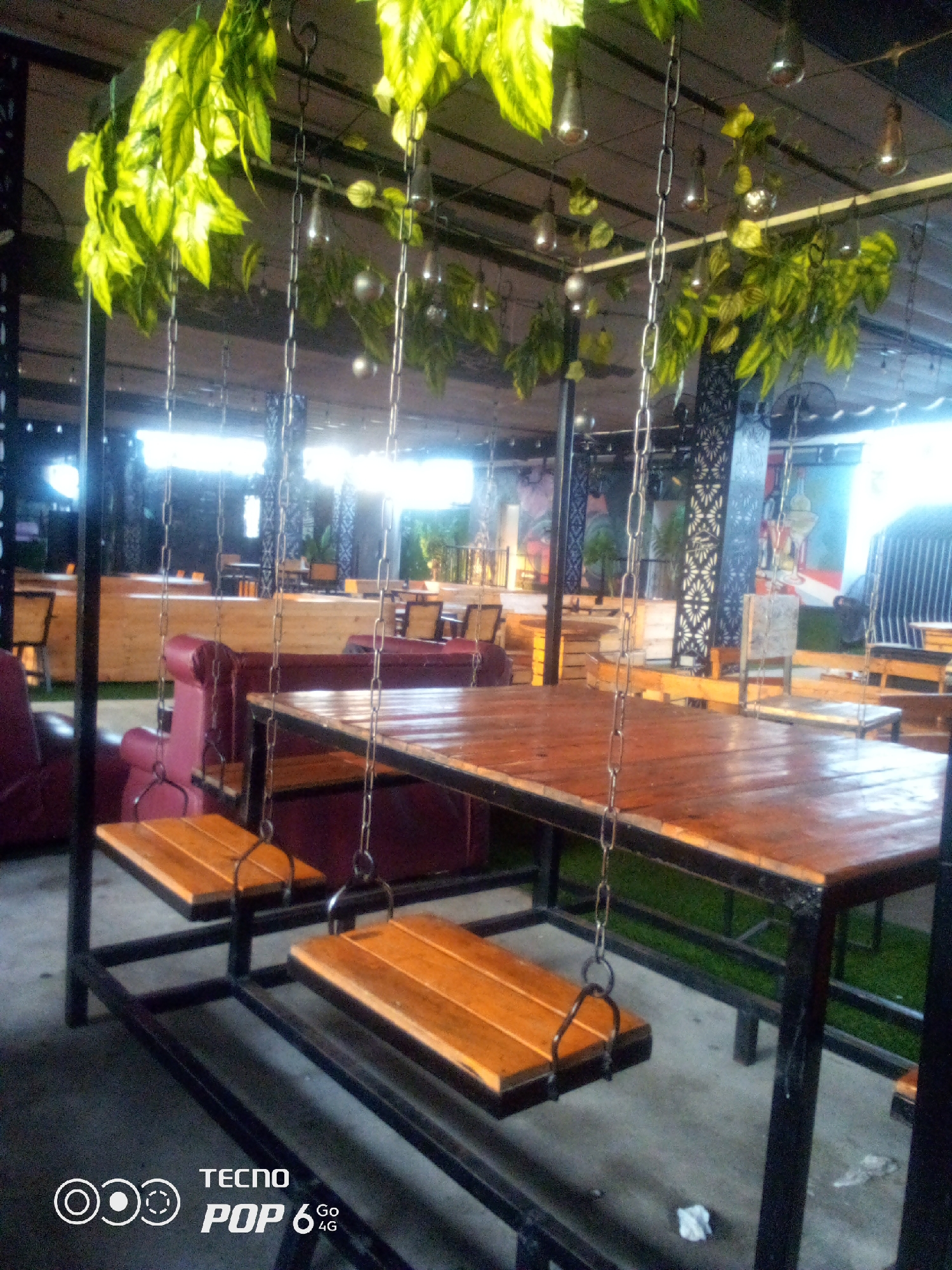 The image captures an outdoor seating area, likely a cafe or restaurant, with a focus on a rustic wooden table supported by a black metal frame. Two wooden swings are suspended by chains from the table's frame, positioned beneath it. Hanging plants with vibrant green leaves and small, reflective light fixtures are visible, suspended from the ceiling structure above. In the background, glimpses of other seating arrangements, including cushioned sofas, wooden benches, and tables, are visible. The setting appears to be in a covered, open-air space, with a bright, diffused light source suggesting it could be daytime or a well-lit evening. The ground surface appears to be a mix of concrete and artificial turf. The bottom right corner of the image contains a "TECNO POP 6 4G" watermark. There are no people visible in the image. This scene is characteristic of outdoor recreational or dining spaces found in urban areas.