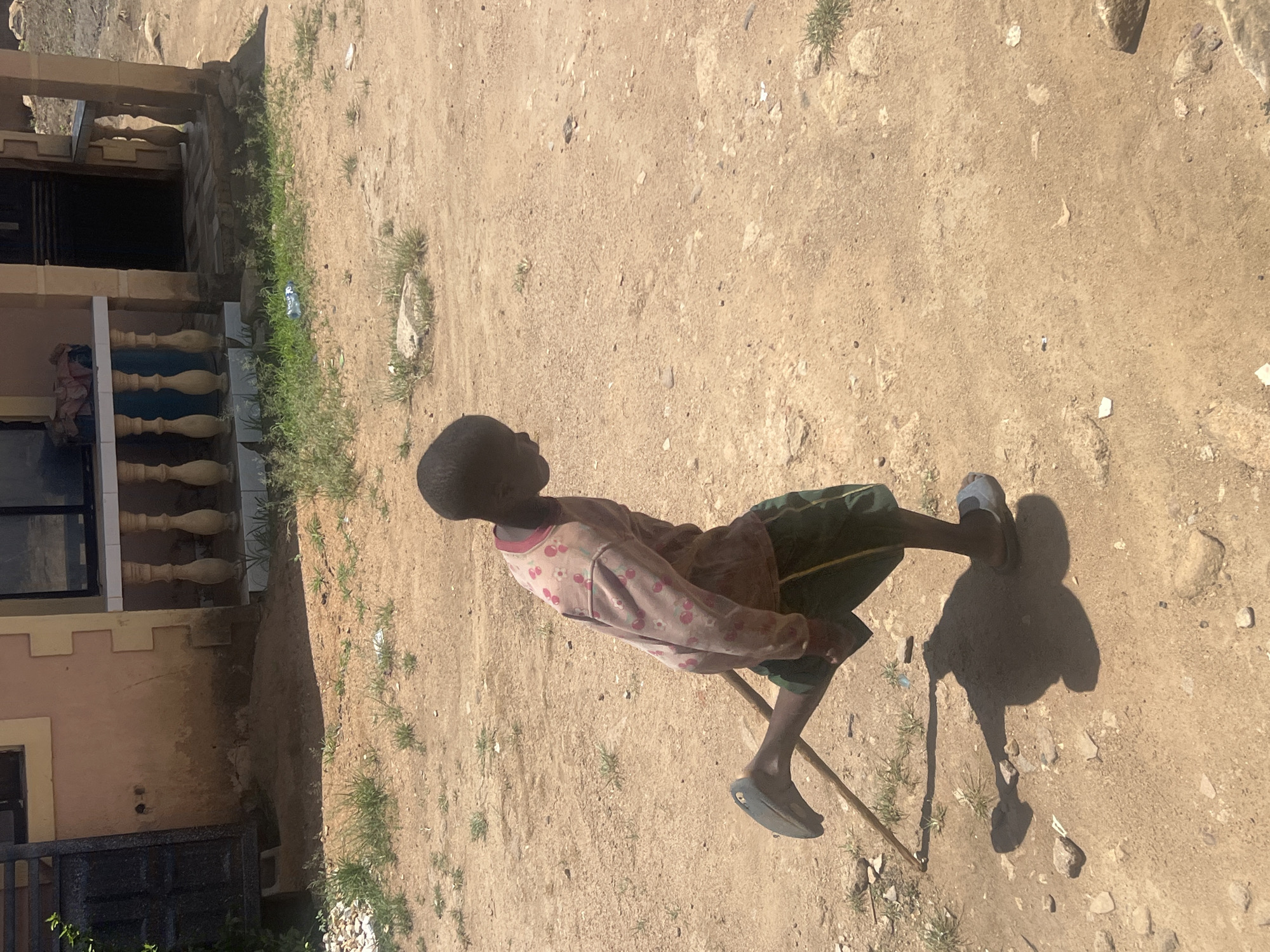 A young person with dark skin sits on a simple wooden chair outdoors in Gura Top, Nigeria. The individual wears a long-sleeved shirt with a floral pattern and green shorts with a yellow stripe, and is wearing one sandal and a single slipper. A stick leans against the chair. The setting is a dusty, dirt-covered yard adjacent to a building with a light-brown exterior, windows, and a balcony with decorative balusters. The ground is bare with sparse patches of grass and some rocks. The scene is bathed in bright sunlight, casting a strong shadow of the subject and the chair onto the ground.