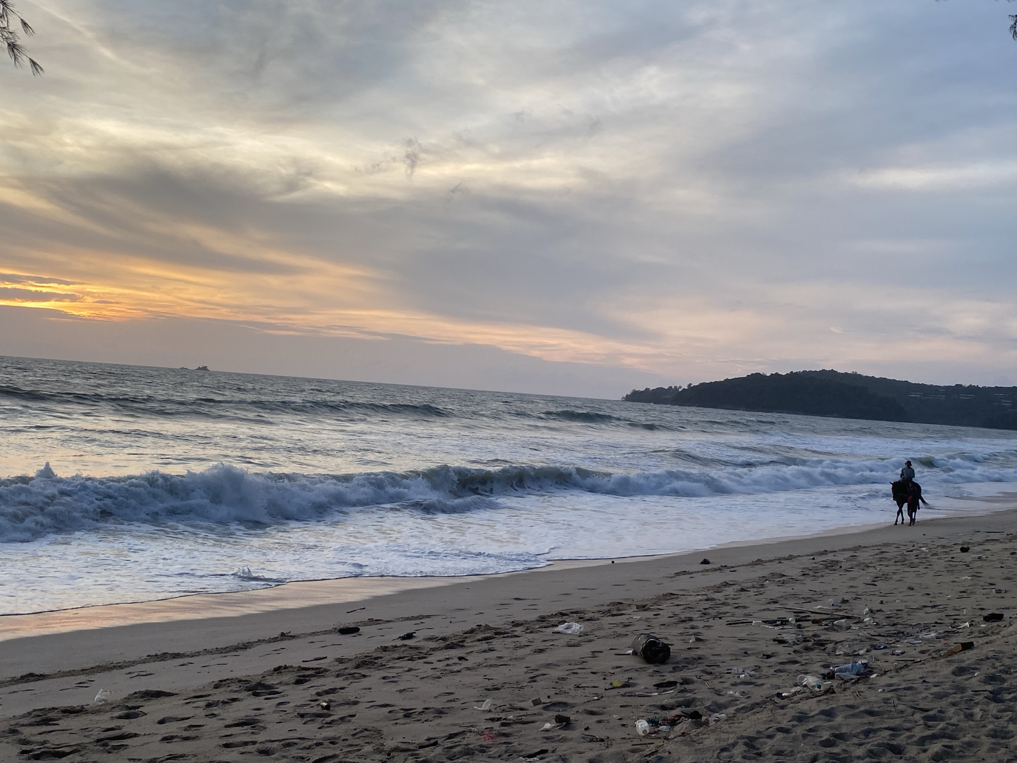 The image depicts a beach in Choeng Thale, Thailand, at dusk. The sky is overcast with shades of grey and pale orange near the horizon, suggesting a sunset. The sea has moderate waves with white foam crests. A figure on horseback is seen riding along the shoreline, toward the right side of the frame. The beach is littered with debris, including plastic bottles and other refuse. In the distance, a dark, hilly landmass is visible.