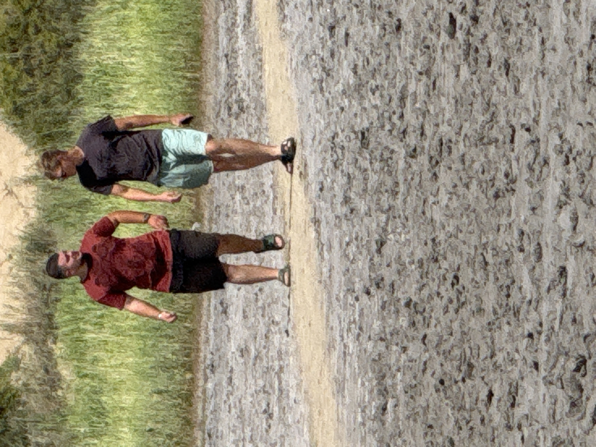 Two men are walking on a dry, cracked, muddy path. The man on the left is wearing a dark red t-shirt and black shorts. The man on the right is wearing a dark gray t-shirt and light blue shorts and is holding a dark object in his right hand. They are walking away from the camera, towards a background of green vegetation and light brown earth. The ground is covered in a textured pattern of dried mud and small rocks. The scene appears to be an outdoor, natural environment. The location is not specified as being in the city of Бурсоль, Russia.