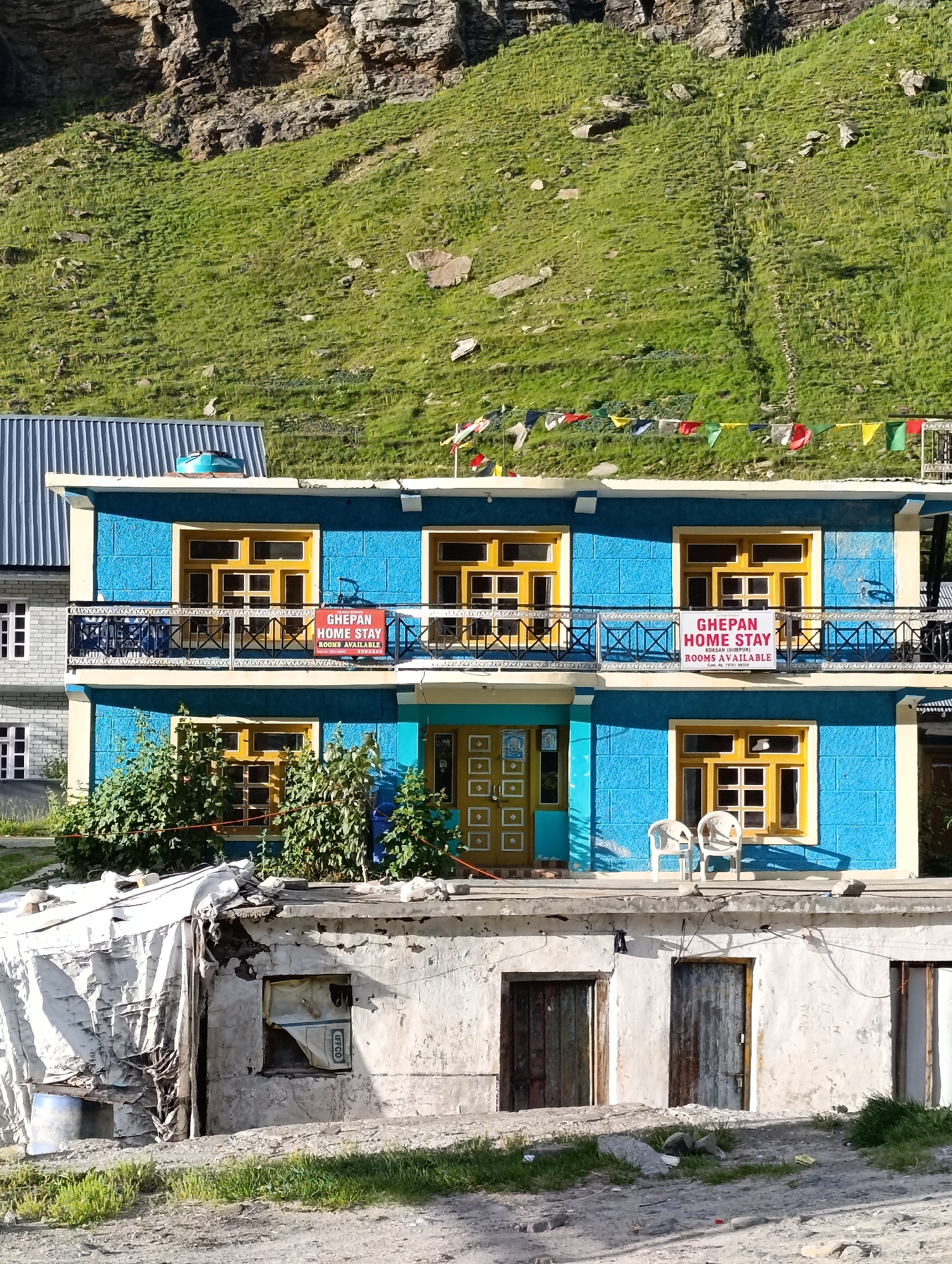 The image depicts a two-story blue and yellow building with a balcony, likely a guesthouse or homestay, situated in the town of Damphug, India, at the base of a grassy, rocky mountainside. The building features yellow-framed windows and a dark railing on the balcony. A red and white sign on the balcony reads "GHEPAN HOME STAY ROOMS AVAILABLE". Prayer flags are strung across the upper section of the building, set against the vibrant green hillside and a rough, rocky cliff face. In front of the building, a lower, weathered structure with concrete walls and worn wooden and metal doors is visible. Two white plastic chairs are placed on the second-story balcony. The foreground shows a rough, unpaved ground with patches of grass and scattered rocks. The scene is bathed in daylight, casting shadows and highlighting the textures of the buildings and natural environment.
