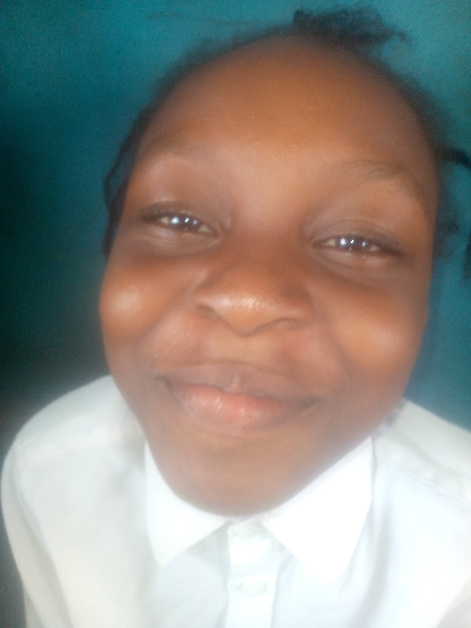 A close-up photograph depicts a Black female with brown eyes, facing the camera with a slight smile. She is wearing a white collared shirt. The background is a solid teal or turquoise color. The location within Ikorodu, Nigeria, cannot be determined based on the visual information provided.