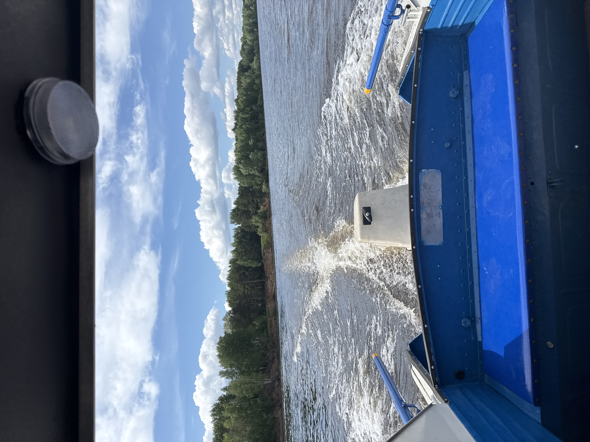 This media file captures a dynamic view from a moving boat on a body of water, likely a river or large lake, during the daytime. The perspective is from inside or from the stern of the boat, looking out towards a distant forested bank and the open sky.
The scene is dominated by the churning water in the immediate foreground and mid-ground, indicating the boat is actively moving. A prominent wake trails behind a white, rectangular object at the back of the boat, which appears to be part of an engine or propulsion system. The boat itself is partially visible, primarily in shades of blue and gray, with visible rivets along its metallic structure. A blue handle or railing extends from the side of the boat. On the far left, a round, translucent light fixture is mounted on a dark, possibly interior, panel of the boat.
In the background, a dense, green forest lines the distant bank of the waterway. Above the treeline, a bright blue sky is adorned with numerous fluffy white cumulus clouds, suggesting clear and sunny weather. Sunlight reflects off the water's surface, creating highlights in the ripples and wake. The time of day is clearly daylight.
There is faint, illegible text on a small, gray label or plate affixed to the blue side of the boat. The image conveys a sense of peaceful travel or recreation on the water. Given the context, this scene is set in Chaikovsky, Russia, potentially on the Kama River or Votkinsk Reservoir.