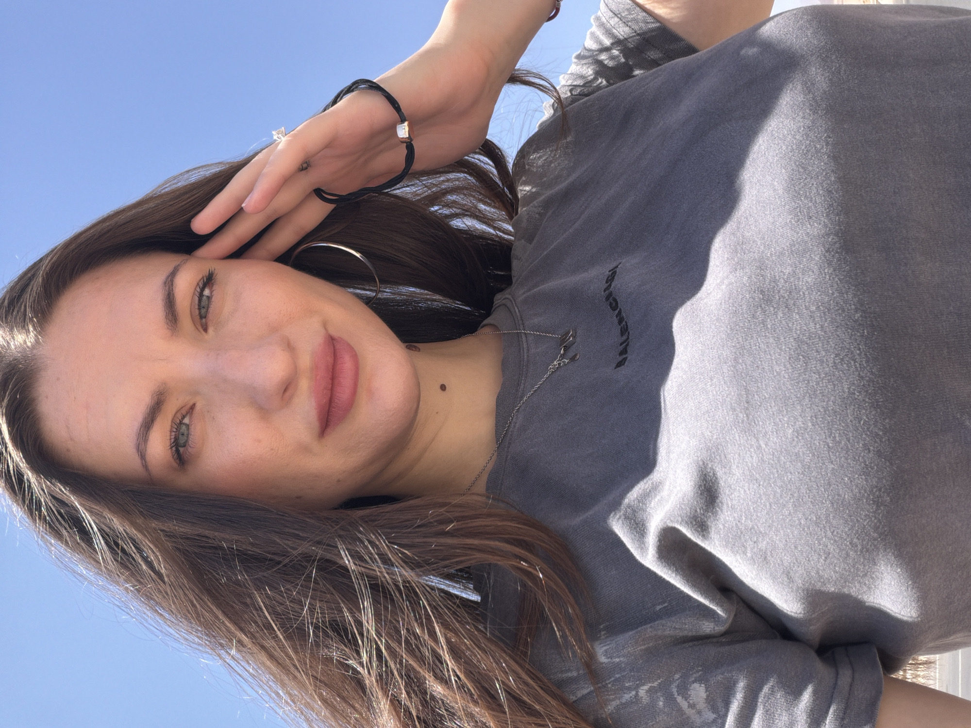 A medium shot selfie shows a young woman with long, brown hair against a clear blue sky. She is wearing a gray t-shirt with a black logo that appears to say "BALENCIAGA". Her left hand is raised, touching her forehead, and she is wearing a ring and a black bracelet with a small gold accent. She also has a large hoop earring in her left ear. A silver necklace with a pendant is visible on her chest. The lighting suggests it is daytime, and the shadows indicate a bright sun. There is no visible indication of the city of Unknown, Egypt, within the image.