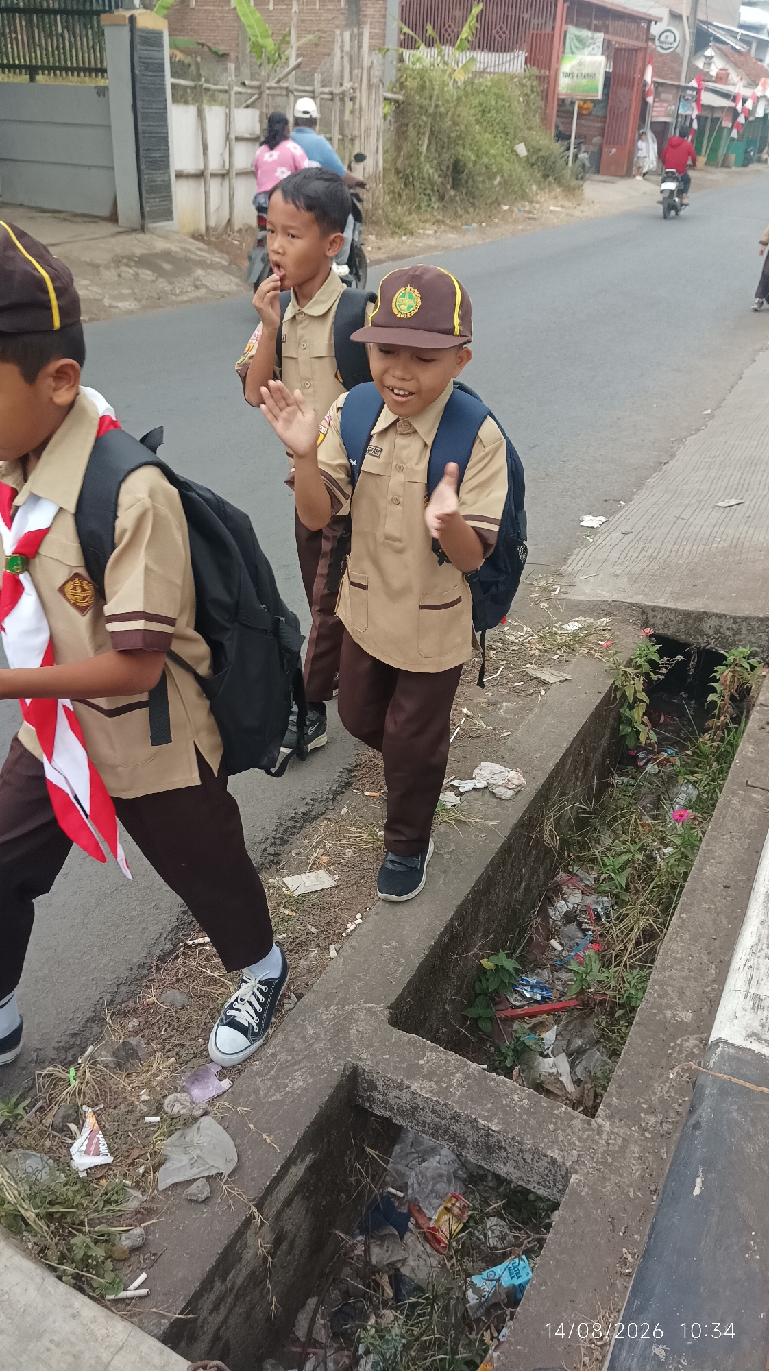 The image captures three young boys in school uniforms, walking along a paved road in Tasikmalaya, Indonesia. They appear to be students, possibly in uniform for scouting activities, given the sash worn by the boy on the left. The boy in the center is wearing a brown cap with a yellow emblem and a brown uniform, with a blue backpack. The boy on the right is also wearing a similar uniform and backpack, with his hands raised as if waving. The road has a concrete drainage ditch on the right side, filled with debris and vegetation, suggesting a less maintained urban area. In the background, there are buildings, some with red and white Indonesian flags, and people on motorcycles, indicating a street scene within a populated area. The overall lighting suggests it is daytime.