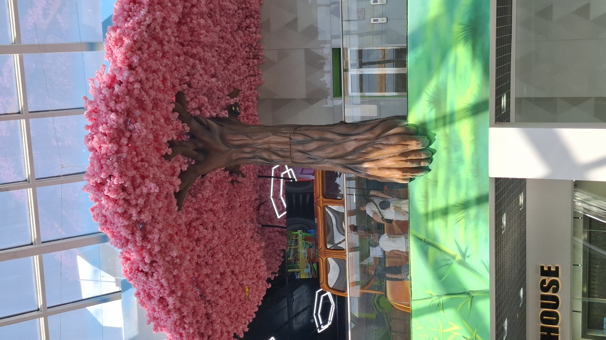 An artificial tree with vibrant pink blossoms is the central focus of this indoor scene, possibly within a shopping mall or entertainment venue. The tree's trunk is detailed and appears to be made of a sculpted material, with its base seemingly emerging from a brightly colored, abstract mural of green foliage. Above the tree, a translucent ceiling with a grid-like structure allows natural light to filter through, illuminating the space. In the midground, a large, orange bus-like structure with tinted windows is visible, and several people, including adults and children, are gathered around it, observing or interacting. Further in the background, there are architectural elements, including glass railings, an elevator, and what appears to be a stage or play area with screens. The overall environment suggests a recreational or commercial indoor space designed for public engagement. The "HOUSE" text visible at the bottom of the image indicates a business or establishment. This setting is consistent with the interior of the Shymkent Plaza shopping and entertainment center in Shymkent, Kazakhstan.