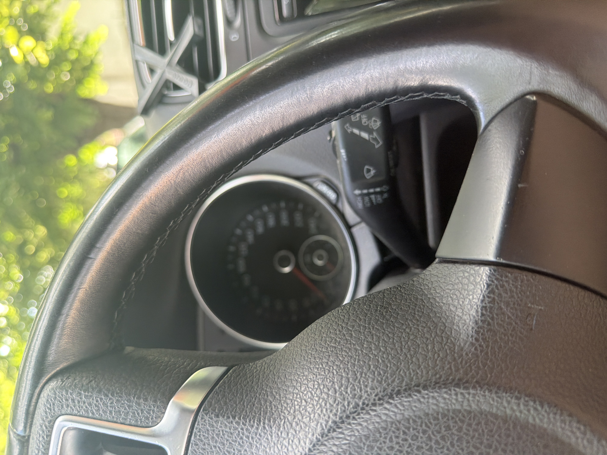 The image captures a close-up view of the interior of a car, specifically focusing on the steering wheel, a portion of the instrument cluster, and a control stalk. The steering wheel, made of black leather with visible stitching along its rim, occupies the foreground and midground, partially obscuring the view of the dashboard. Some wear and tear are visible on the leather surface of the steering wheel.
Through the upper gap of the steering wheel, a round instrument dial, likely a speedometer, is visible. Its needle rests near the zero mark, suggesting the car is stationary. To the right of the instrument cluster, a black control stalk is clearly visible, featuring icons for wiper settings, including "OFF" and various speeds/modes for front and rear wipers.
In the upper-left background, outside the car, bright, blurred green foliage and sunlight create a bokeh effect, indicating daytime and an outdoor setting. Above the steering wheel and to the left, a car air vent is visible, partially showing a stylized symbol that resembles an "X" or a winged "A", with some faint, unreadable text below it. The car appears to be located in Iskar, Bulgaria.
