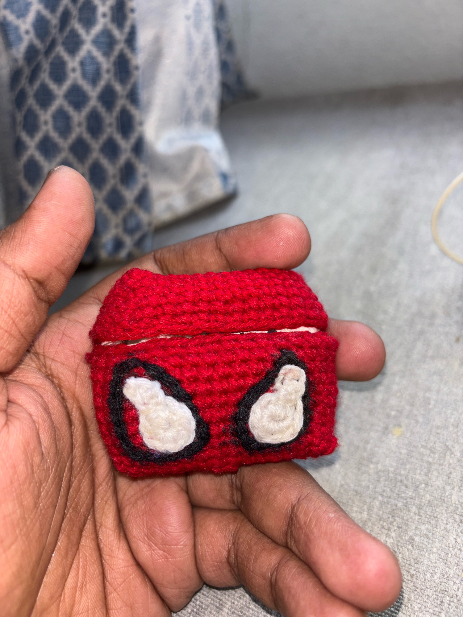 A person's hands are holding a red crocheted object that resembles a Spiderman mask. The object is small and rectangular, with two white oval shapes outlined in black, representing Spiderman's eyes. The background is blurred and shows a patterned fabric and a gray surface. The image is taken from a close-up perspective, focusing on the object and the hands. The lighting is natural, casting soft shadows. There is no information available to determine the location within Ghataprabha, India.