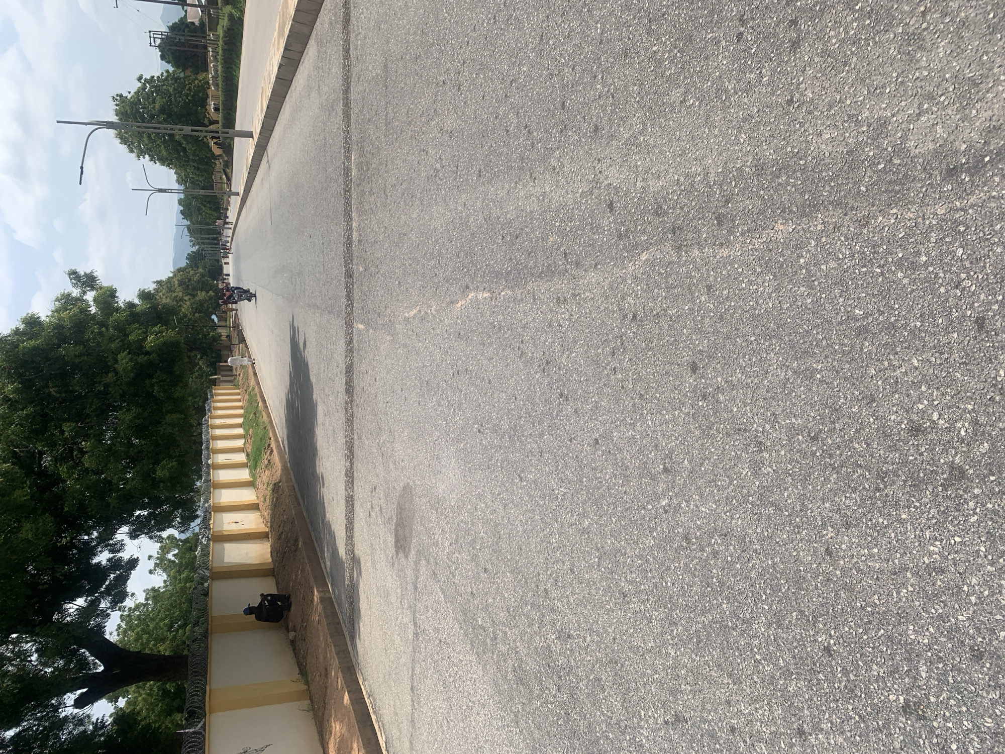 The image depicts a street in Jalingo, Nigeria. The asphalt road stretches into the distance, flanked by trees and a yellow and white wall with barbed wire. A person in dark clothing and a blue cap is seated by the wall on the left side of the road. Further down the road, several people and a motorcycle are visible. Streetlights are intermittently positioned along the right side of the road, which has a concrete curb and shoulder. The sky is partly cloudy.