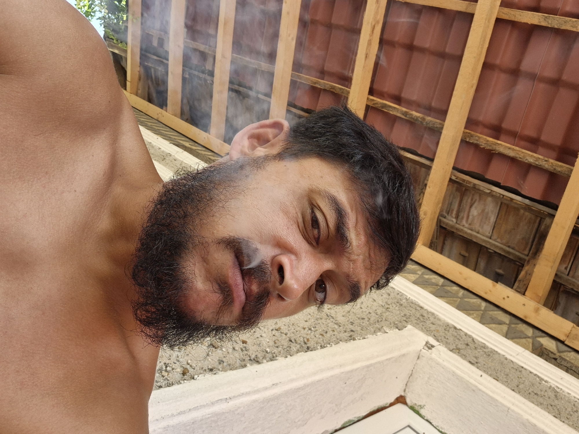A close-up, low-angle selfie shows a shirtless man with dark hair and a full beard smoking. He is outdoors, with a wooden roof structure and a tiled roof visible in the upper left background. To the right, a textured wall with decorative tiling and white trim is present. The man's face is turned slightly towards the camera, with smoke emanating from his mouth. The background suggests a rustic or rural setting, possibly in Drianovets, Bulgaria, given the traditional roofing materials and exterior wall construction. The lighting indicates it is daytime, with sunlight illuminating the scene.