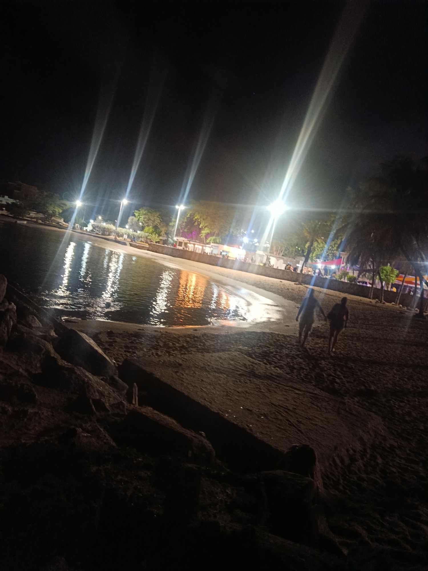 The image depicts a beach scene at night in Juan Griego, Venezuela. Several streetlights illuminate the beach and the calm water, creating reflections on the surface. Two individuals, appearing to be a couple, are walking hand-in-hand on the wet sand near the water's edge. In the background, there are palm trees and structures, suggesting a developed coastal area with amenities. The foreground shows rocks and sand, characteristic of a beach environment. The lighting creates a dramatic and somewhat hazy atmosphere, with light beams from the streetlights stretching across the sky.