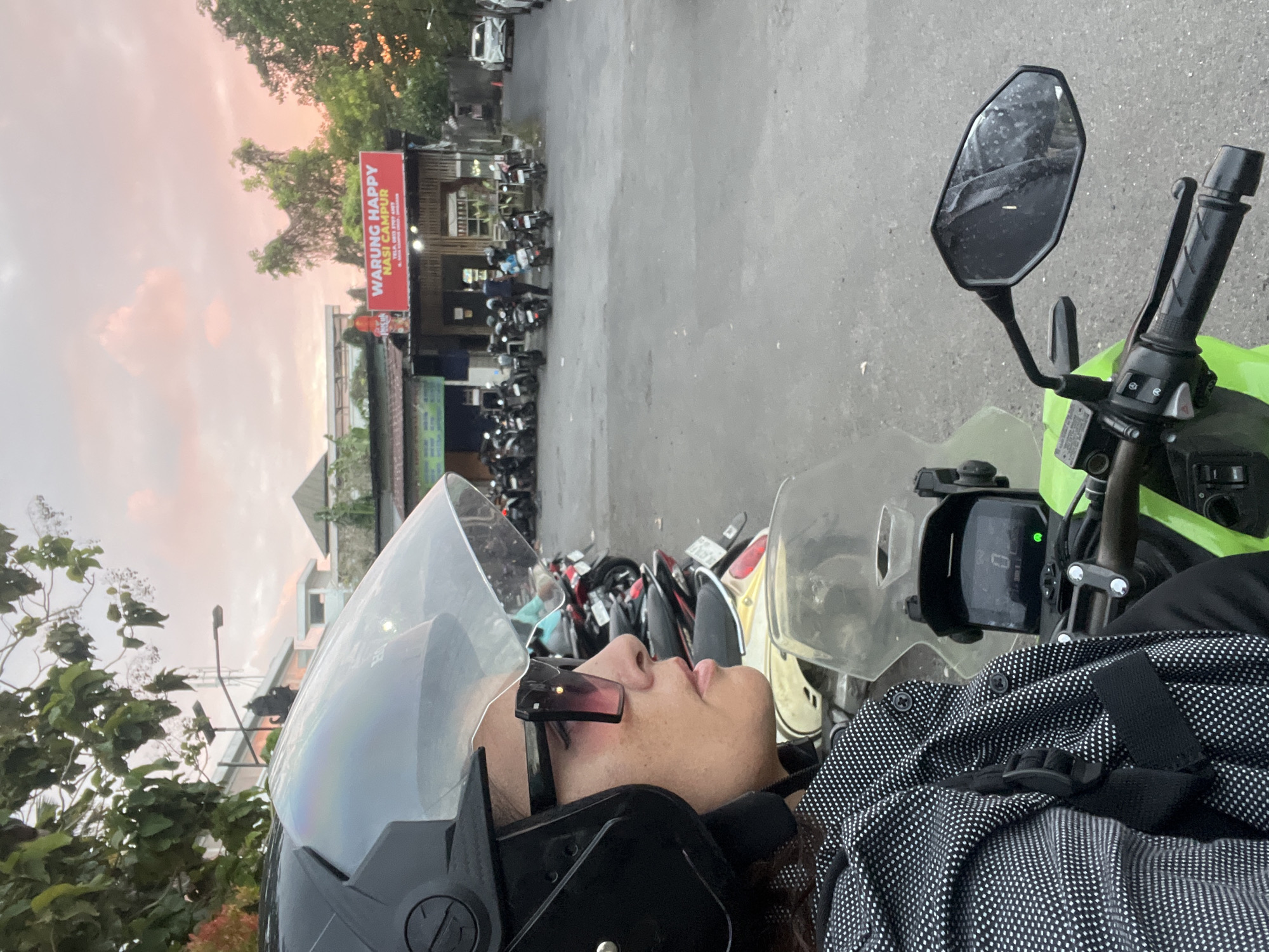 In Kuta Selatan, Indonesia, a woman wearing a black helmet and large sunglasses is seated on a green motorcycle. She is facing to the right, with her profile visible. Her clothing appears to be a black and white patterned jacket. The motorcycle's dashboard shows a digital display with the number "0". In the background, a "WARUNG HAPPY NASI CAMPUR" sign is visible above a building. Numerous other motorcycles are parked in the area. The sky above is filled with soft pink and orange clouds, suggesting either sunrise or sunset. The scene is set on an asphalt surface, likely a street or parking lot, with trees and other buildings forming the urban environment.