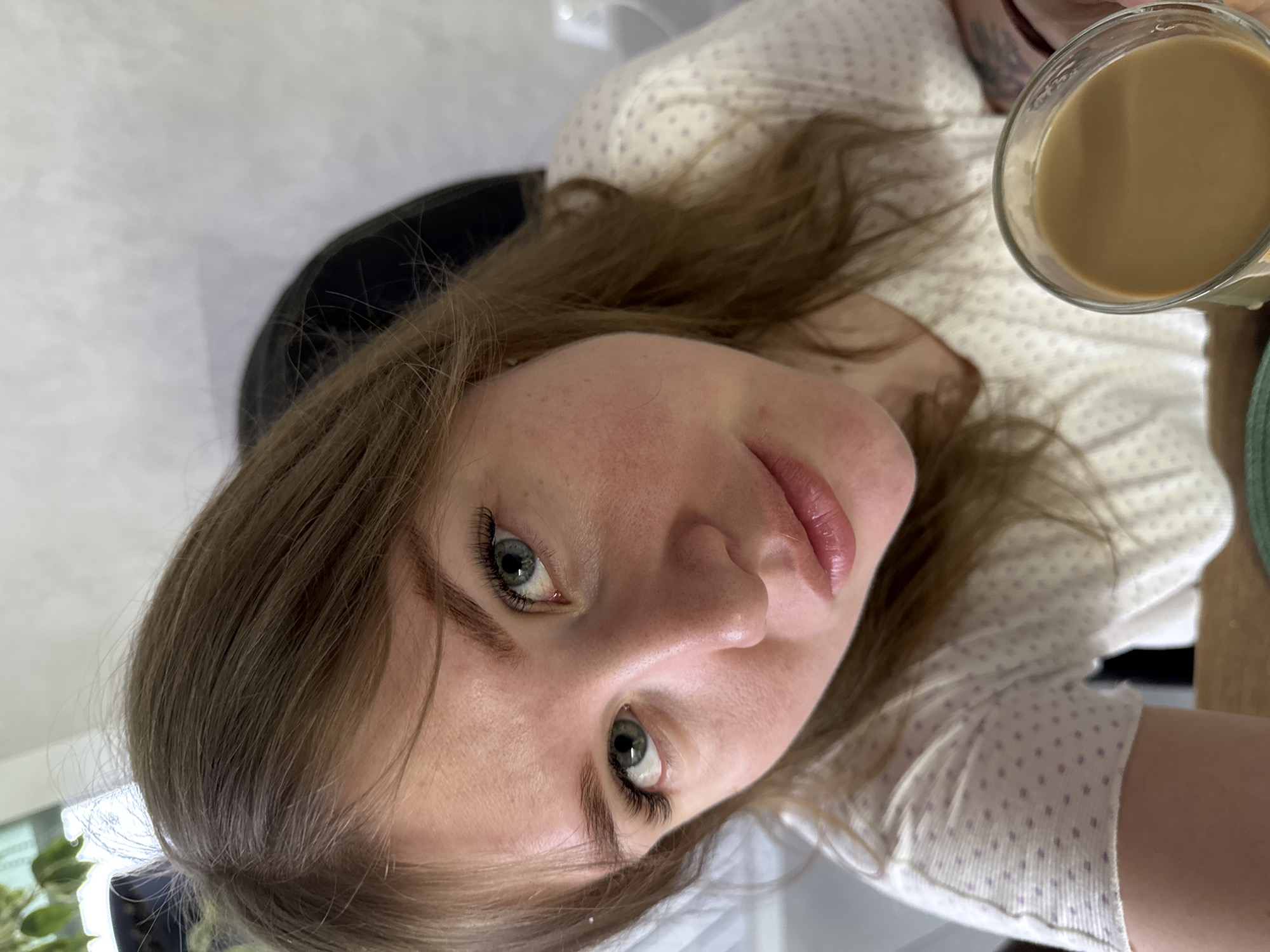 The image depicts an indoor scene in Gornyi (Dvurechenskoe S/P), Russia. A young adult female with light brown hair and prominent blue-green eyes gazes directly into the camera. She wears a light-colored, short-sleeved top adorned with small, light purple polka dots. Her right hand, visible in the lower right quadrant, holds a clear glass filled with a light brown, opaque liquid, consistent with coffee. A dark-colored tattoo is partially visible on her left forearm/wrist area. The background features out-of-focus light-colored walls, a dark object possibly an office chair behind her, and green foliage of a houseplant in the upper left, suggesting proximity to a window. The lighting is soft and even.