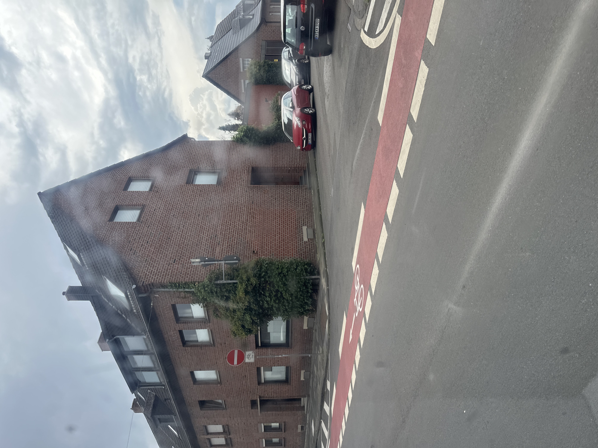 The image depicts a street scene in Krefeld, Germany, likely on a cloudy day. In the foreground, a red-painted bicycle lane with white dashed markings is visible on the asphalt road. To the left, a multi-story brick building with several windows and a prominent pitched roof dominates the scene. A red and white circular "no entry" sign with a bicycle symbol is affixed to a pole on the sidewalk in front of this building. Further down the street, a red hatchback car and two other vehicles are parked. In the background, another brick building with a pitched roof is visible, suggesting a residential or mixed-use urban area. The overall impression is of a quiet street with typical German architecture and urban amenities.