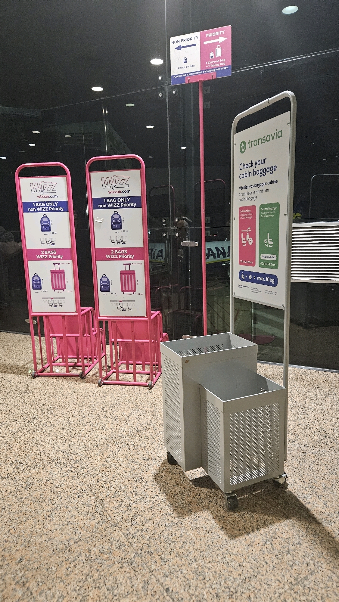 The image depicts a brightly lit indoor area, likely an airport terminal, with a patterned tiled floor. Two pink metal frames, each displaying information about Wizz Air baggage policies, are positioned side-by-side. These frames are supported by rolling bases. To the right, a taller, white metal stand with a sign for Transavia airline is visible. This sign details cabin baggage allowances and has a small, gray wheeled bin attached to its base. The bin has two compartments and a perforated metal design. In the background, reflective glass panels reveal a dimly lit interior with some indistinct figures and another sign with the word "ANA" visible. The context strongly suggests this scene is within Barcelona-El Prat Airport in El Prat de Llobregat, Spain, given the presence of airport signage and baggage check information for airlines operating in such a facility.