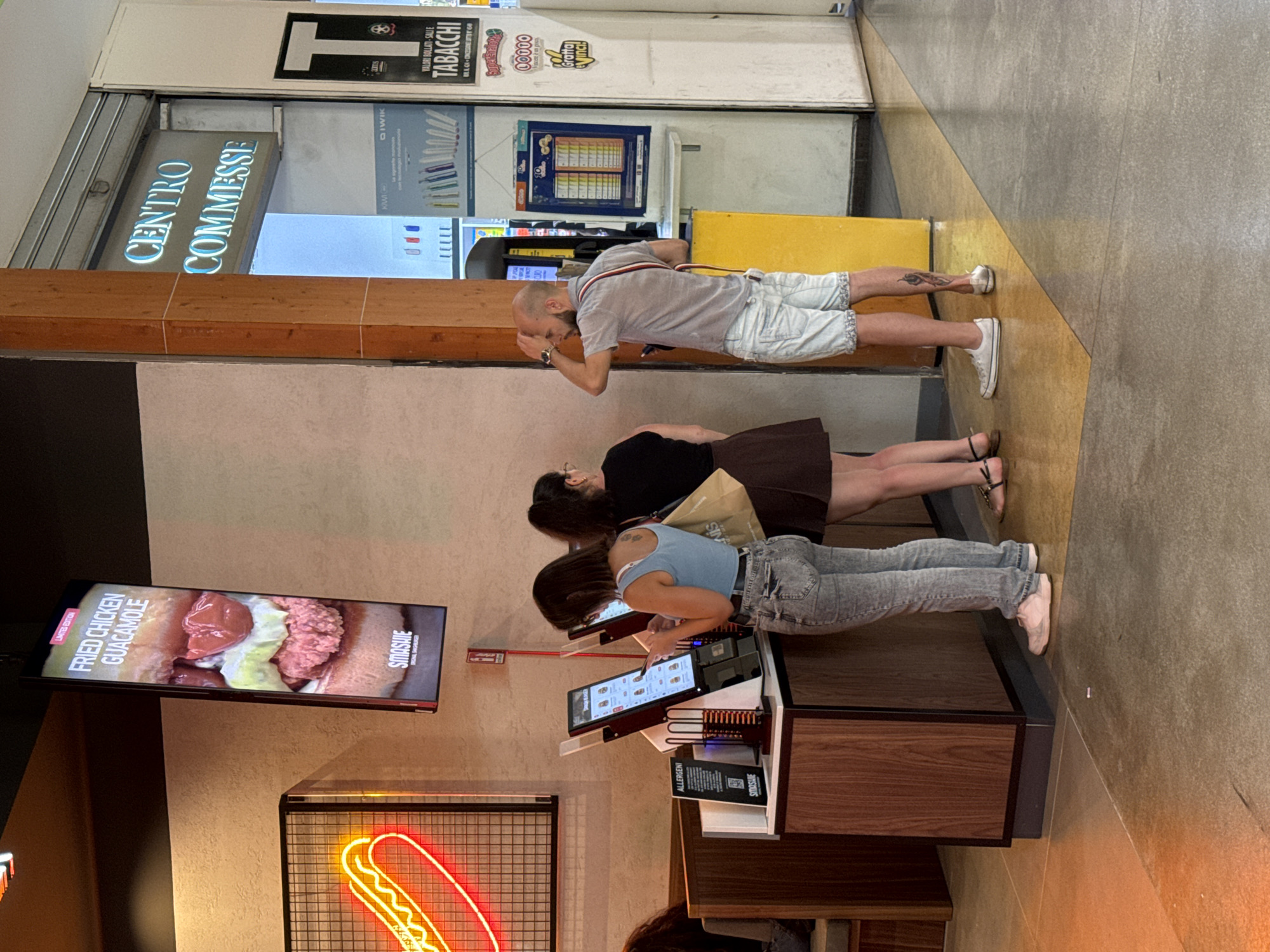 The image depicts an interior scene, possibly a fast-food establishment or a shopping center with food vendors, in Palermo, Italy. On the left, a digital menu board displays an advertisement for "FRIED CHICKEN GUACAMOLE" from a brand named "SMASHIE ORIGINAL BURGER". Adjacent to it is a neon sign shaped like a hot dog. In the center, two women and one man are standing near self-ordering kiosks. One woman, wearing a blue tank top and grey jeans, is actively using a kiosk. Another woman, in a brown skirt and black top, stands behind her, and the man, in a grey t-shirt and white shorts, is looking to his right. The background reveals a sign that reads "CENTRO COMMESSE" (Shopping Center) in illuminated blue letters and various advertisements for tobacco products and lotteries. The flooring is a mix of polished concrete and yellow tiles. The overall impression is a busy commercial space with modern ordering technology.