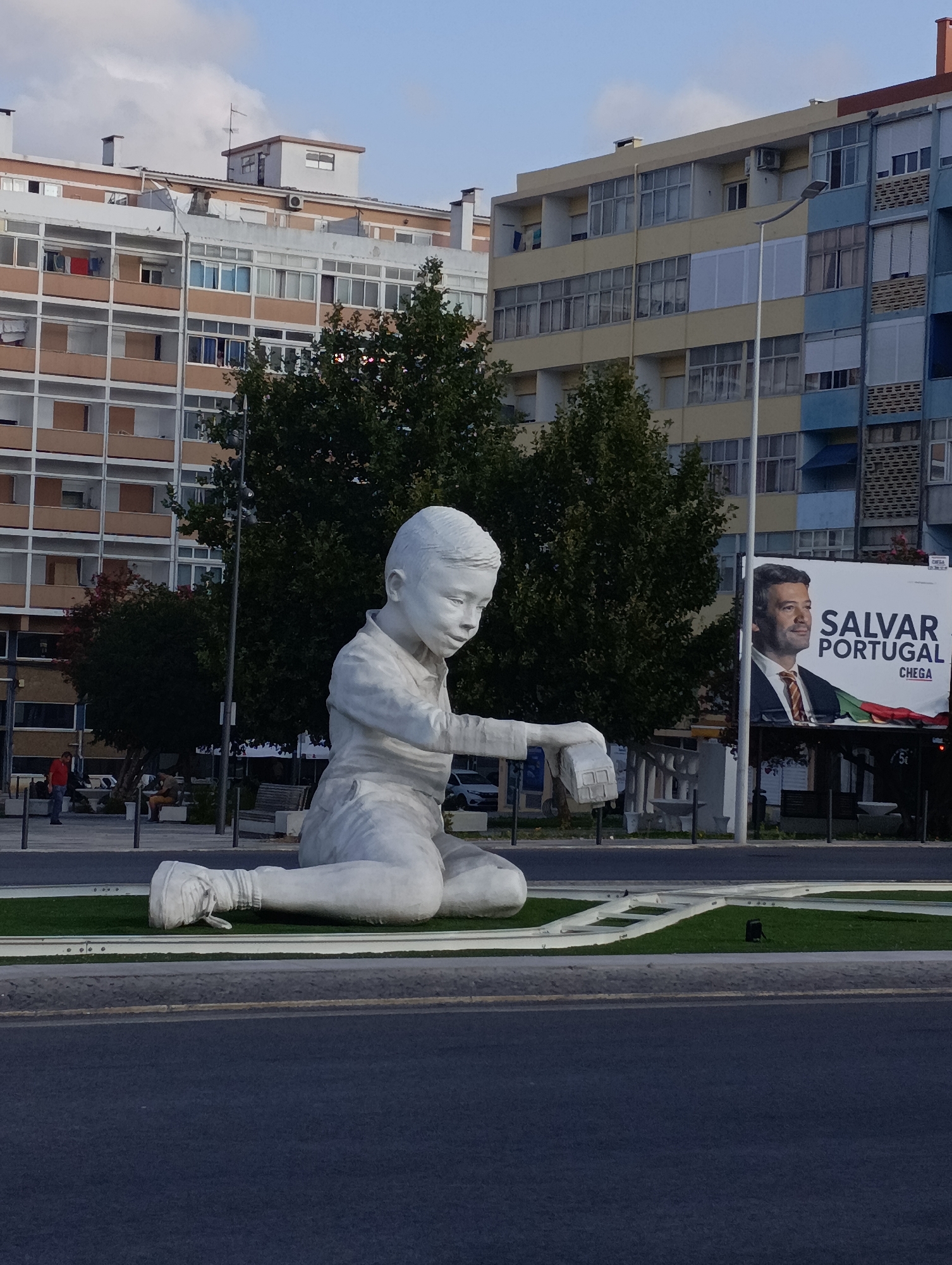 A large, white sculpture of a young boy, depicted in a seated, contemplative pose, dominates the foreground of this outdoor scene. The boy's left arm is extended, holding a small, rectangular object. He is wearing sneakers, one of which is partially visible and appears to be white with laces. The sculpture rests on a grassy area surrounded by a paved walkway and road. In the background, several multi-story apartment buildings with various colored facades, balconies, and numerous windows are visible. Trees, some with dark green foliage and one with reddish leaves, are situated between the sculpture and the buildings. A large billboard featuring a man's portrait and Portuguese text that reads "SALVAR PORTUGAL CHEGA" is also present in the background. The sky above is a pale blue with scattered white clouds. The overall setting appears to be a public urban space in Setúbal, Portugal, possibly a roundabout or a plaza, indicated by the presence of the sculpture, billboard, and apartment buildings. Several individuals are sparsely distributed in the background, appearing to be pedestrians and possibly sitting on benches.