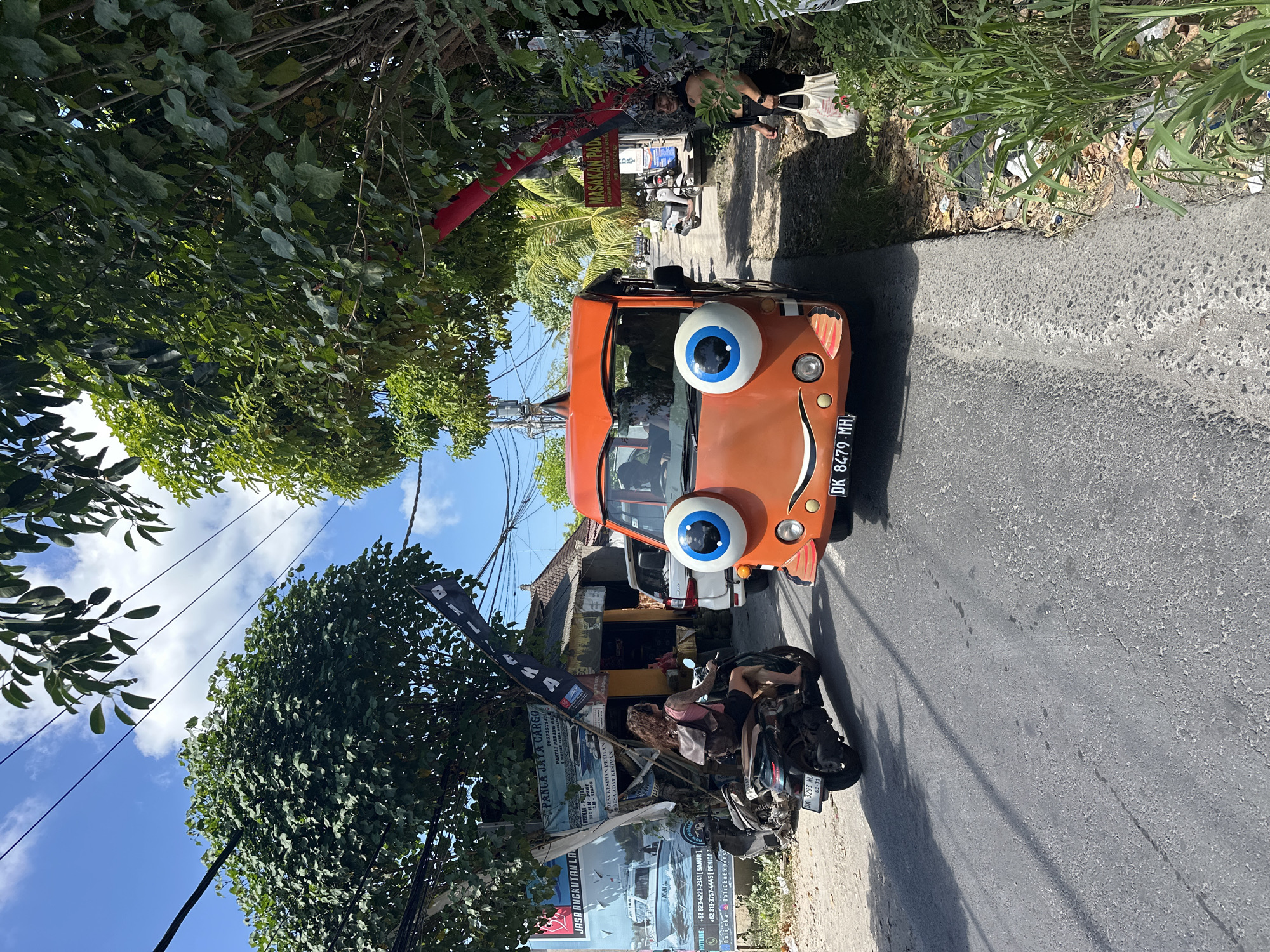 The image depicts a narrow road in Nusa Penida, Indonesia, flanked by tropical vegetation and buildings. A distinctive orange vehicle with large cartoon eyes attached to its hood dominates the foreground. Parked alongside the road near the vehicle is a motorcycle with a large advertising banner attached to it. The banner promotes "PANCA JAYA CARGO" and provides contact information, including phone numbers for Sanur and Penida. Other text on the banner includes "JASA ANGKUTAN LA" and "DESA KESIMAN PETILAN DESA ADAT KESIMAN". Another sign is visible for "MASAKAN PAD" which could be a food establishment. At the edge of the road is trash/vegetation. A person is seen standing near a building further up the road. The sky is visible through gaps in the foliage. The vehicle license plate reads "DK 8479 MH". Another license plate on the motorcycle is seen as "DK 32G3 NL."