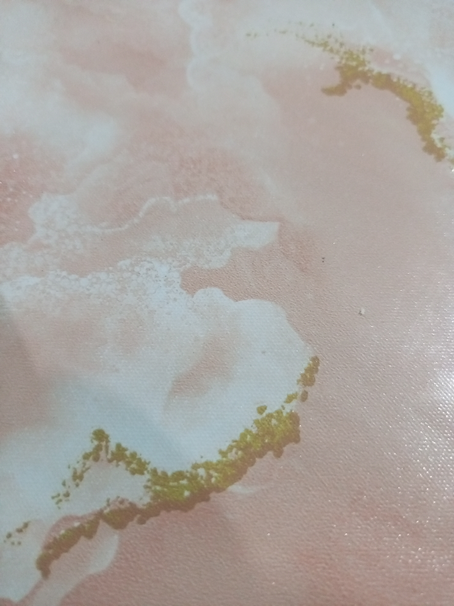 The image displays a close-up, abstract view of a textured surface, possibly fabric or a painted canvas, with a predominantly pale pink and white marbling effect. Interspersed within this marbling are streaks of shimmering gold particles, forming irregular patterns. There are no people, objects, actions, or interactions visible. The scene is not located in the city of Lhokseumawe, Indonesia, as the image lacks any identifiable geographical or contextual markers.