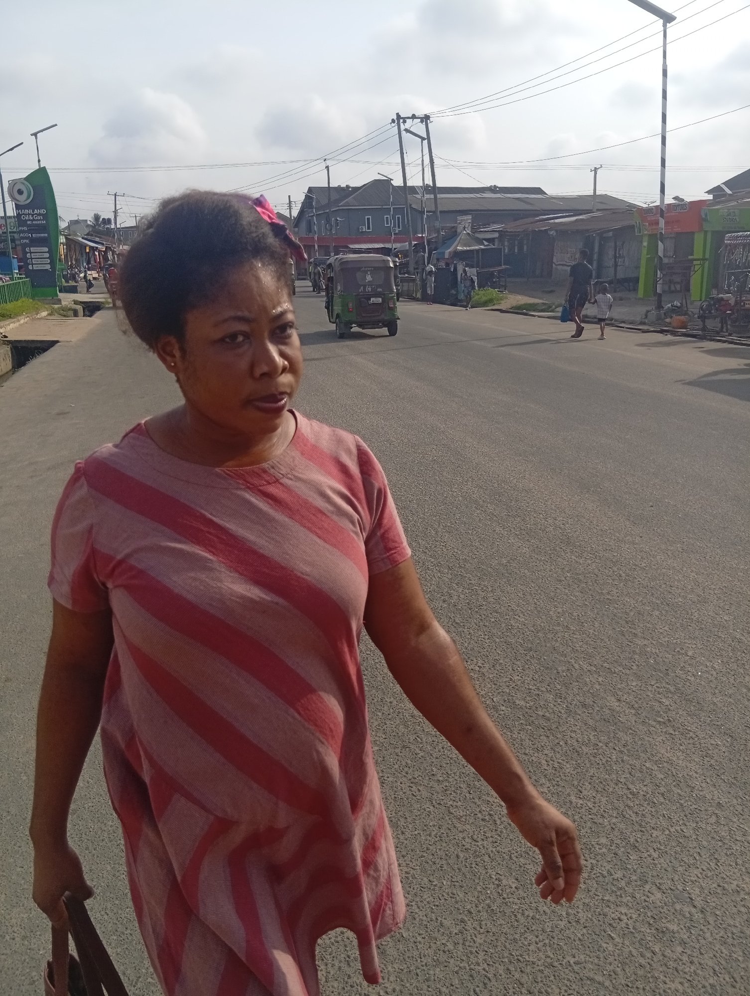 A woman is walking down a paved street in Aba, Nigeria. She has dark hair with a pink hair accessory and is wearing a striped, knee-length dress. In the background, there are buildings, utility poles, and other people. A green tricycle-type vehicle is in the street. Other vehicles are parked near the buildings. The sky is overcast.