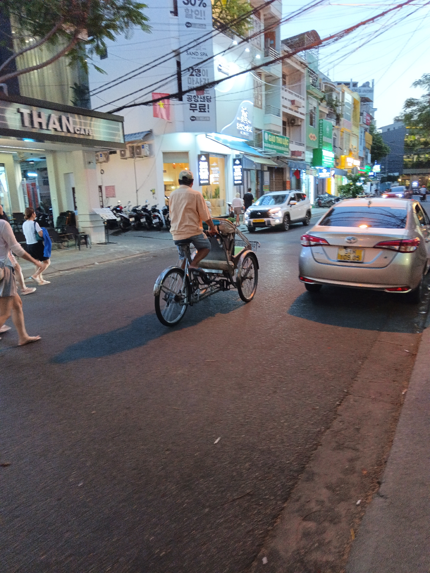 This is an eye-level, medium shot of a street scene in Nha Trang, Vietnam, likely taken at dusk or dawn due to the lighting conditions. In the foreground, a person wearing a peach-colored shirt and grey shorts is riding a three-wheeled pedicab, facing away from the viewer. The pedicab has a metal frame and a covered passenger area behind the rider.
To the left of the pedicab, three women are walking on the sidewalk. The woman closest to the viewer is wearing a light pink dress and a straw hat. The middle woman is wearing a white t-shirt and a black skirt, holding a blue bag. The third woman, partially obscured, is wearing a white t-shirt and a black skirt.
The background features a row of multi-story buildings with various signs and awnings, indicative of commercial establishments like shops and cafes. One prominent sign reads "THAN CAFE". The street is paved and appears to be a busy urban area with overhead power lines crisscrossing the sky. Several motorcycles are parked along the sidewalk in front of one of the buildings.
A silver sedan with a yellow license plate is driving on the street, slightly behind the pedicab. Another white SUV is also visible further down the street. The overall atmosphere suggests a typical street scene in a Vietnamese city, with a mix of pedestrians, vehicles, and commercial activity.