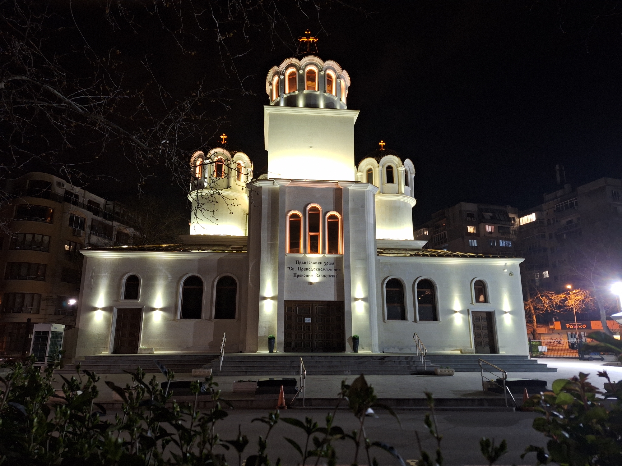 The image captures the Saint Procopius of Varna Orthodox Church in Varna, Bulgaria at night. The church is predominantly white and well-lit, with three dome-shaped structures topped with crosses. The central, tallest dome is tiered. Each dome features arched windows that are illuminated from within. The church's facade has multiple arched windows and a set of double doors at the center, flanked by spotlights. A sign in Cyrillic script is above the central doorway. Steps lead up to the church entrance. The foreground includes greenery, which partially obscures the view. The background is blurred but shows apartment buildings on either side of the church. Bare tree branches are also visible on the left side. The lighting is artificial, enhancing the church's architectural details.