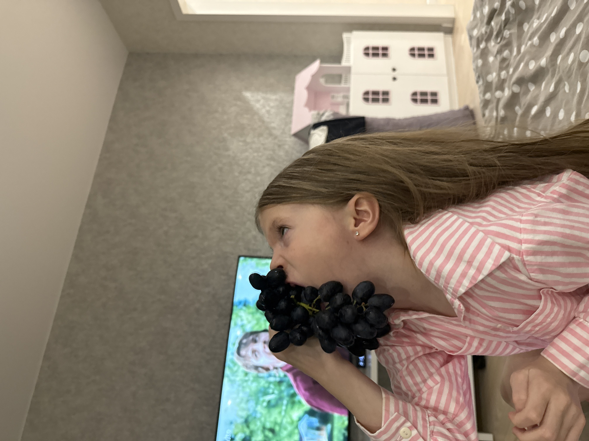 A young girl with long, light brown hair, wearing a pink and white striped shirt, is holding a bunch of dark purple grapes to her mouth, appearing to eat them. She is in a room with light gray walls and a white ceiling. A television screen is visible in the background, displaying an image of people outdoors, possibly from a show or movie. Behind the girl, on a piece of furniture, there is a white dollhouse with pink accents. A polka-dot patterned blanket is partially visible on what appears to be a bed. The location is likely indoors and there is no information to verify the location of Izhevsk, Russia.
