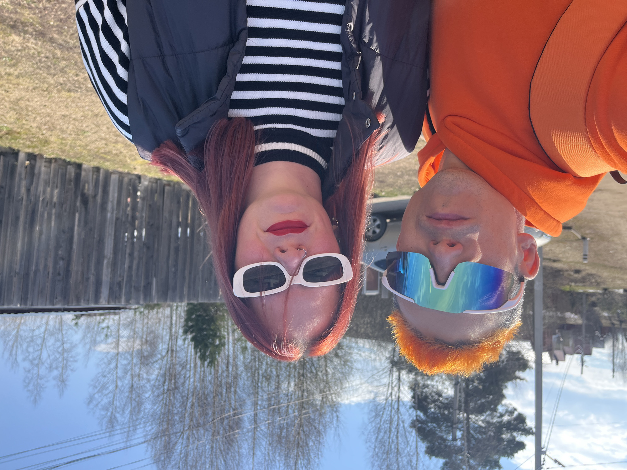 Two individuals, a woman and a man, are captured in a bright outdoor portrait in Chaikovsky, Russia. The image is taken on a sunny day, likely in late autumn or early spring, given the bare trees in the background.
The woman, positioned on the left, has long reddish-brown hair. She wears white, rectangular-framed sunglasses, red lipstick, and a nose ring. Her attire consists of a black and white horizontally striped long-sleeve top layered under a black puffer vest.
The man, on the right, has vibrant orange hair and sports blue and green mirrored sports sunglasses. He is wearing a bright orange hoodie. Both individuals are looking directly towards the camera, posing for the shot.
The background features a rustic wooden fence behind the woman, extending horizontally across the frame. Beyond the subjects, numerous bare trees are visible against a clear blue sky. A light-colored car is partially visible in the mid-ground. Power lines can also be seen in the distance against the sky. The bright sunlight casts clear illumination on the subjects and their surroundings. There is no visible text in the image.