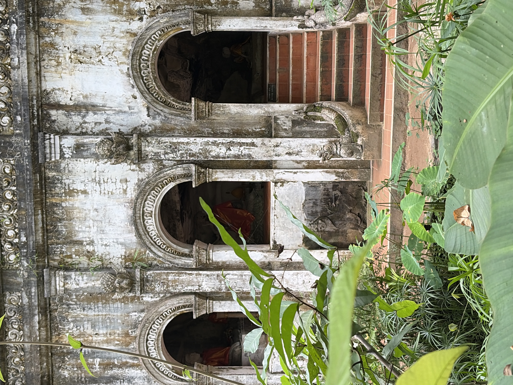 The image shows an exterior view of an ancient temple structure in Suthep Town Municipality, Thailand. The weathered white stone facade features three arched alcoves, each containing a seated Buddha statue draped in orange cloth. The central and right alcoves are partially obscured by lush green foliage in the foreground. The architecture exhibits intricate decorative carvings along the top of the facade and around the arches. Stone dragon-like figures guard the base of a set of red-tiled stairs leading into the rightmost alcove. The ground in front of the structure is covered with reddish-brown soil and more dense vegetation.