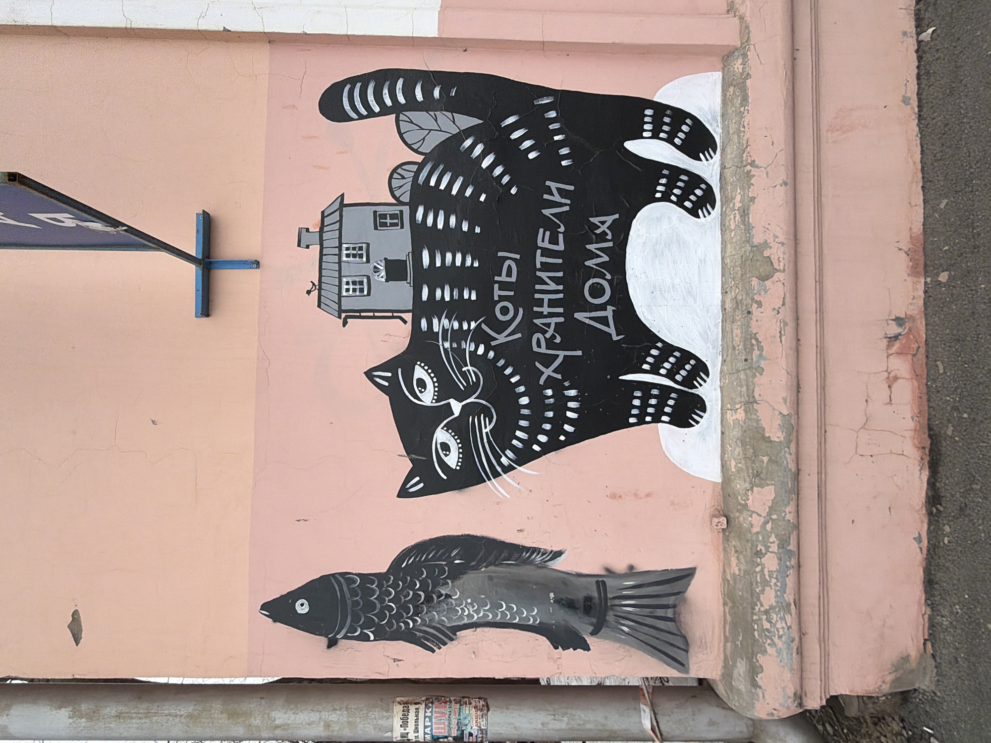 This is an outdoor shot of a wall mural in Votkinsk, Russia. The mural is on a light pink, textured wall that also has a darker pink decorative trim along the right edge. The mural consists of a large, black cat with white stripes and markings, facing to the left. On top of the cat is a small grey house with white windows. To the right of the cat, written in white Cyrillic script, is "Коты Хранители Дома" (Cats Guardians of the House). Below the cat is a black fish with grey markings.
Other details include a blue sign and its metal attachment on the left side of the wall and a horizontal, grey pipe near the bottom of the frame. There is also some trash and debris along the bottom. The trim has some peeling paint. The wall is uneven, and the paint is cracked in places. The right edge of the shot is bordered by a section of asphalt.