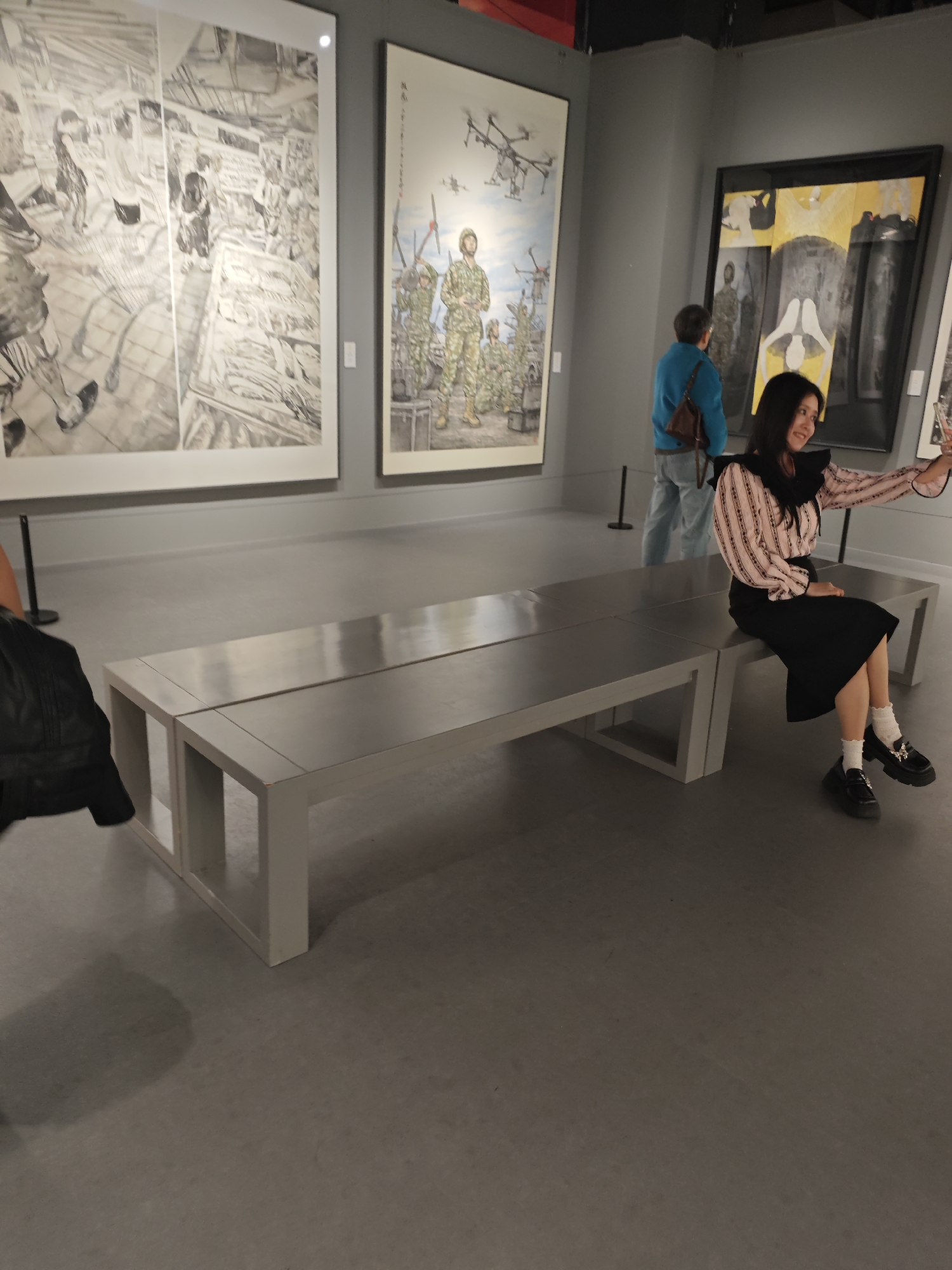 A woman is sitting on a long metal bench in an art gallery, taking a selfie with her phone. The gallery has multiple large paintings on the walls, one of which shows soldiers and drones. Another person stands in the background. The floor is gray. A portion of a black jacket is visible in the bottom left of the image.