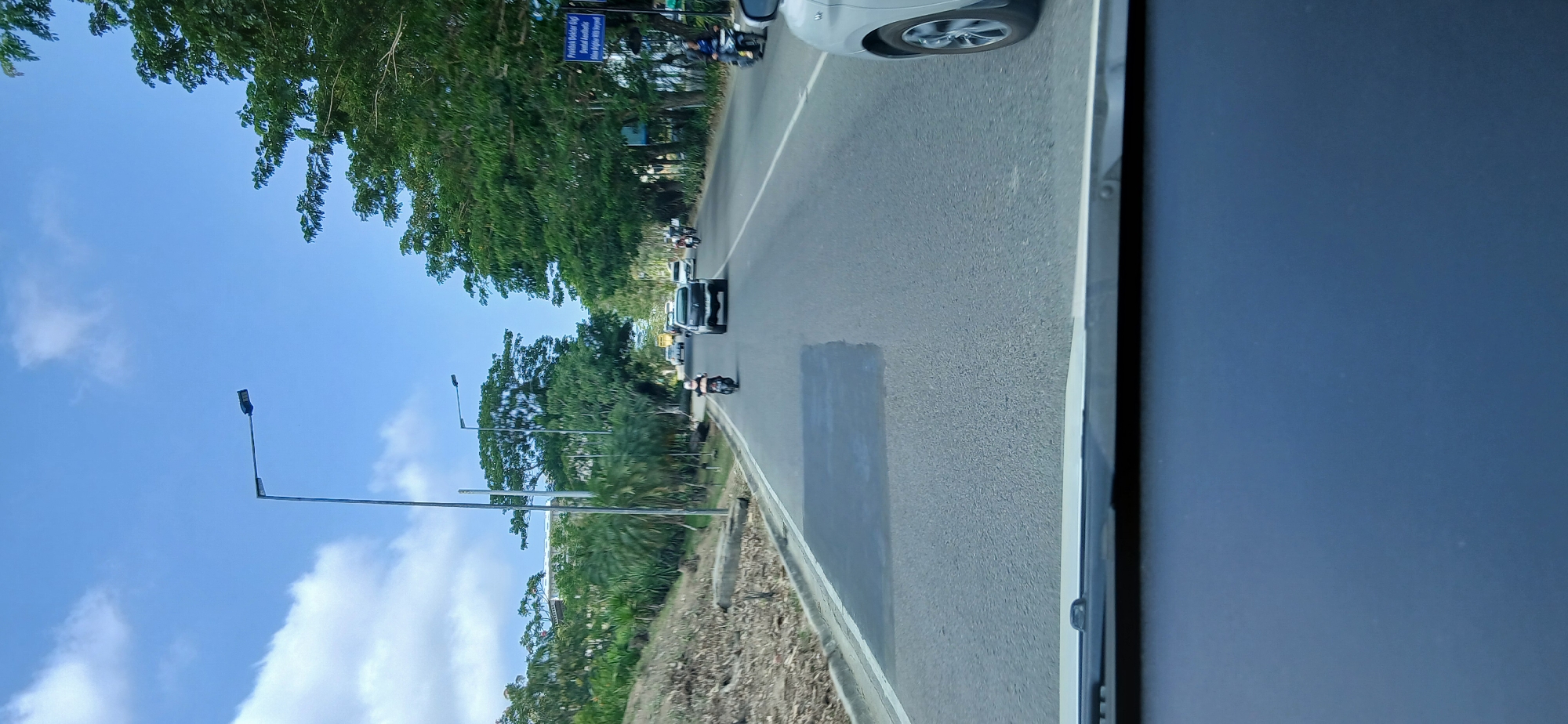 The image shows a view from inside a vehicle looking out at a road in the city of Samarinda, Indonesia. The road is asphalt and appears to be a multi-lane highway. There are several vehicles on the road, including a motorcycle, a black SUV, and a silver car. Trees line the right side of the road, and there is a small hill with vegetation on the left side. The sky is blue with some white clouds. A street light is visible on the left side of the road. A sign is visible on the right side of the road with text that is difficult to read but appears to be in Indonesian. The overall scene suggests a sunny day with moderate traffic.