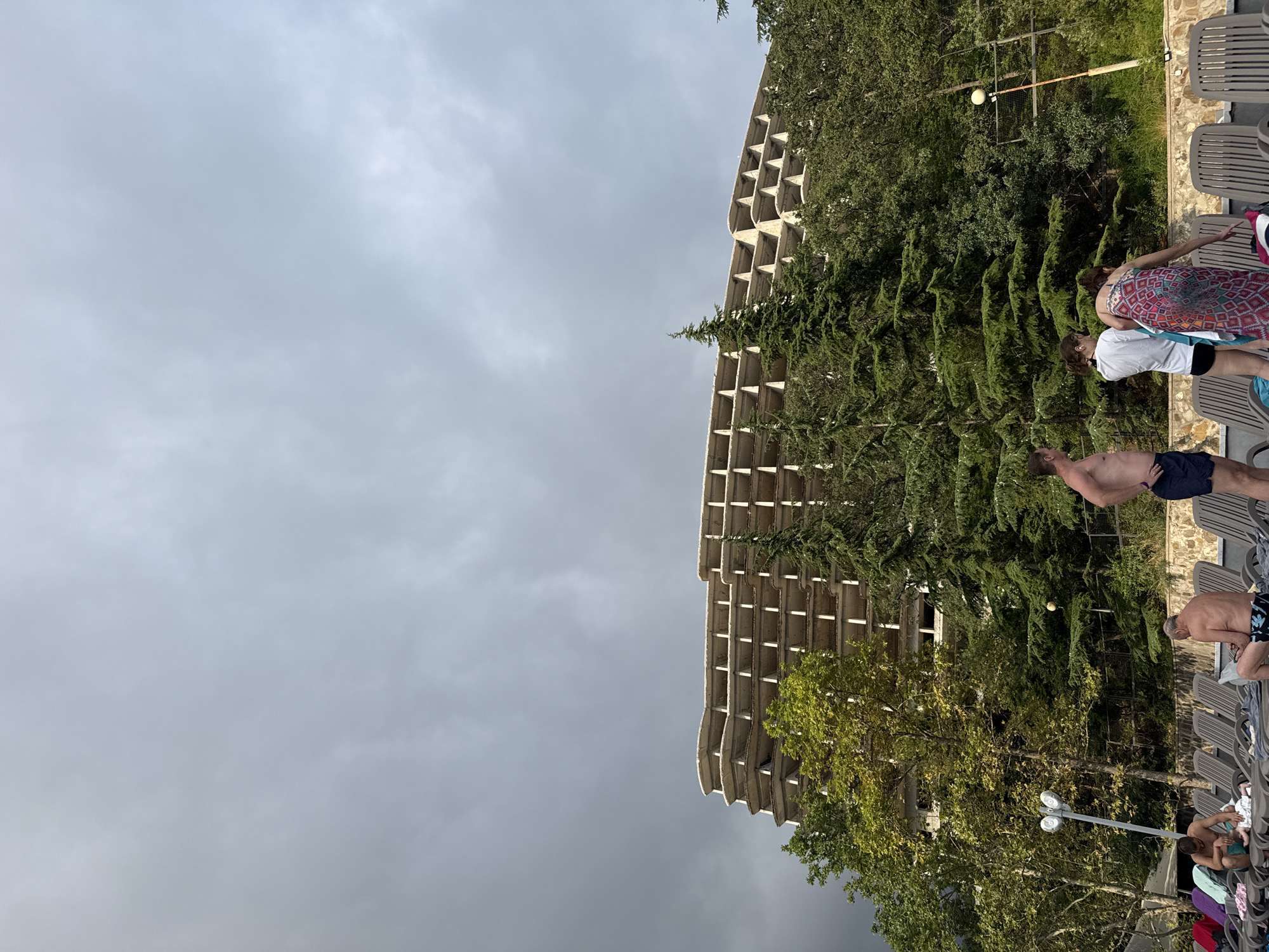 The image depicts a somewhat overcast day with dark clouds dominating the sky. In the background, a large, unfinished concrete building with multiple balconies and exposed structural elements stands prominent. It appears to be derelict or under construction, surrounded by dense green foliage, including trees that partially obscure its lower levels. In the foreground, a group of people are visible near a row of empty lounge chairs, suggesting a resort or public area. One man is standing with his back to the camera, wearing swim trunks. Another man, also in swim trunks, is seated on a lounge chair. A third person, wearing a patterned sarong and a white top, is partially turned away, with two other individuals standing nearby. The overall scene suggests a recreational area set against the backdrop of a large, incomplete structure.