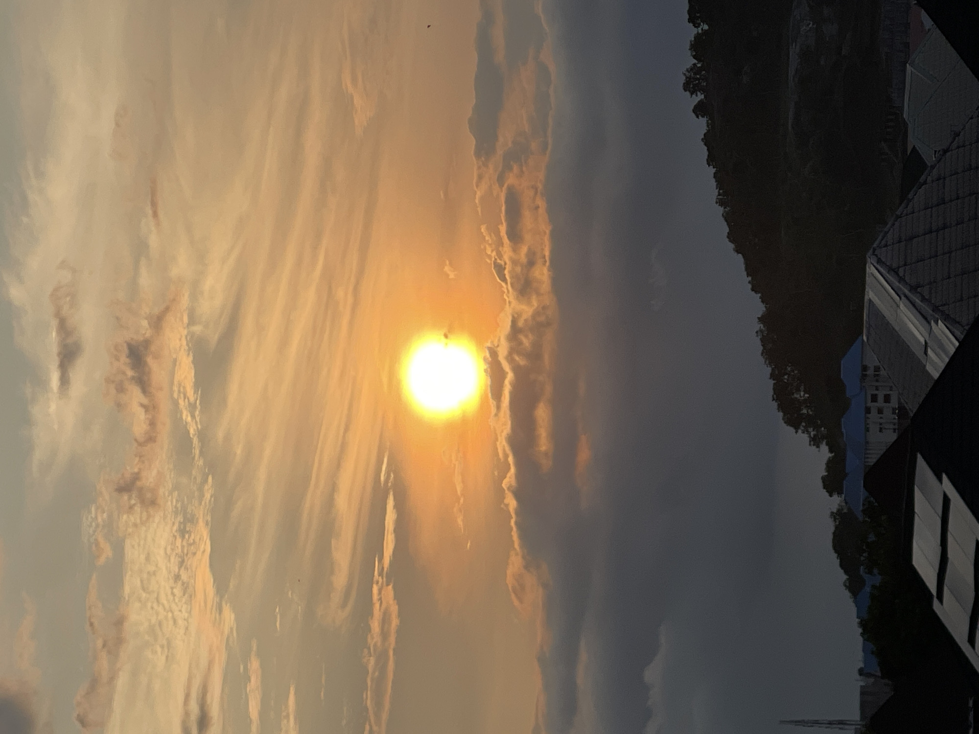 The image captures a sunset over the city of Abuja, Nigeria. The sun, a bright orange orb, is partially obscured by wispy clouds, casting a warm glow across the sky. The clouds are a mix of white and light orange, with darker, more textured clouds below. In the foreground, silhouetted against the sky, are the rooftops of buildings, some with distinct tiled patterns. Beyond the rooftops, the dark outline of a hill covered in trees rises, with glimpses of buildings and a tower in the distance. The overall scene is one of natural beauty as the day transitions into evening.