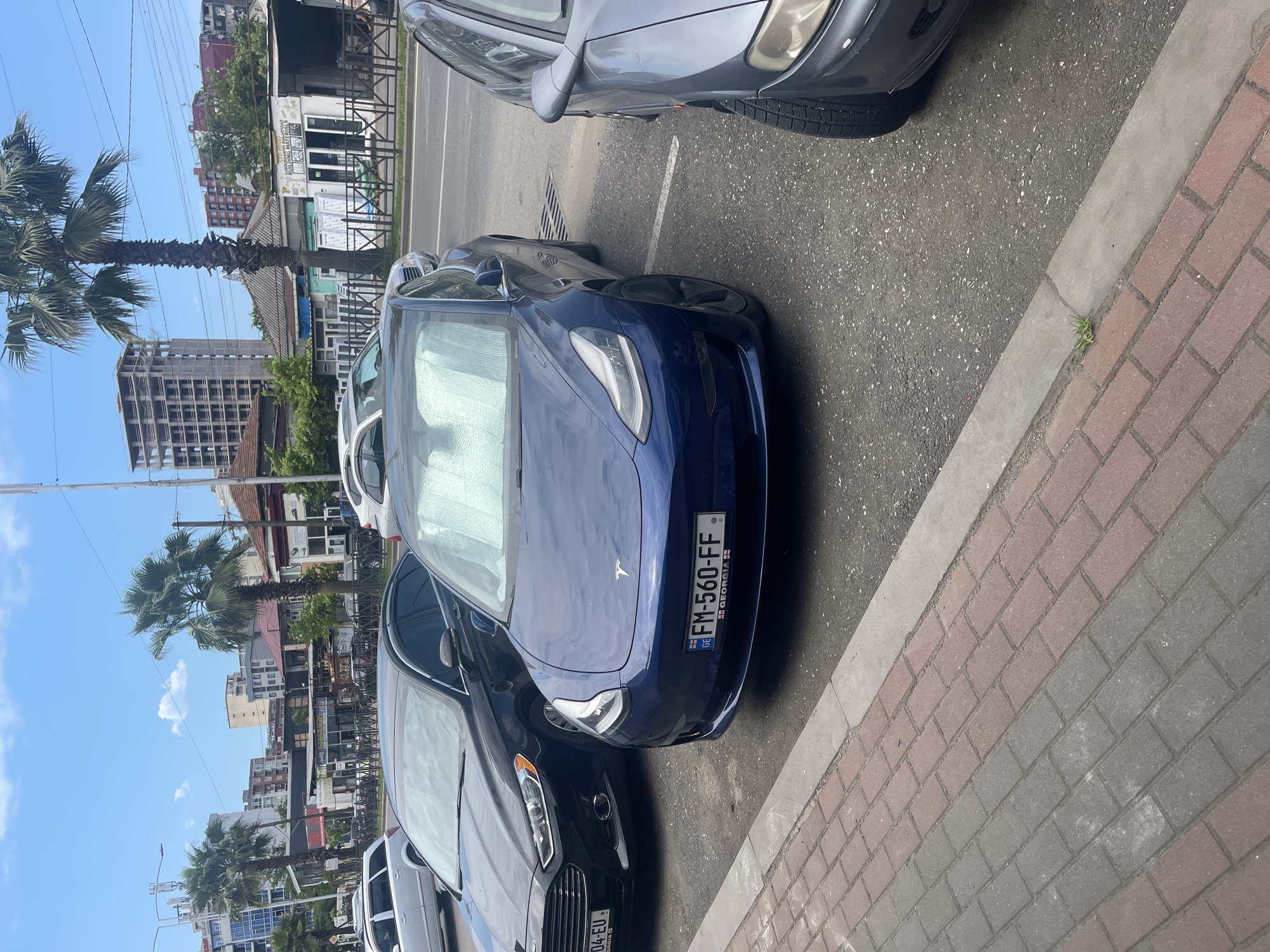 The image shows a row of parked cars on a street in Batumi, Georgia. The foreground is dominated by the hood of a blue Tesla, with its license plate reading "FM-560-FF GEORGIA". To the left of the Tesla is a dark-colored car, and further back, a silver car. In the background, buildings and palm trees line the street. The sky is blue with scattered white clouds, suggesting it is daytime and the weather is clear. The scene depicts a typical urban streetscape with parked vehicles.
