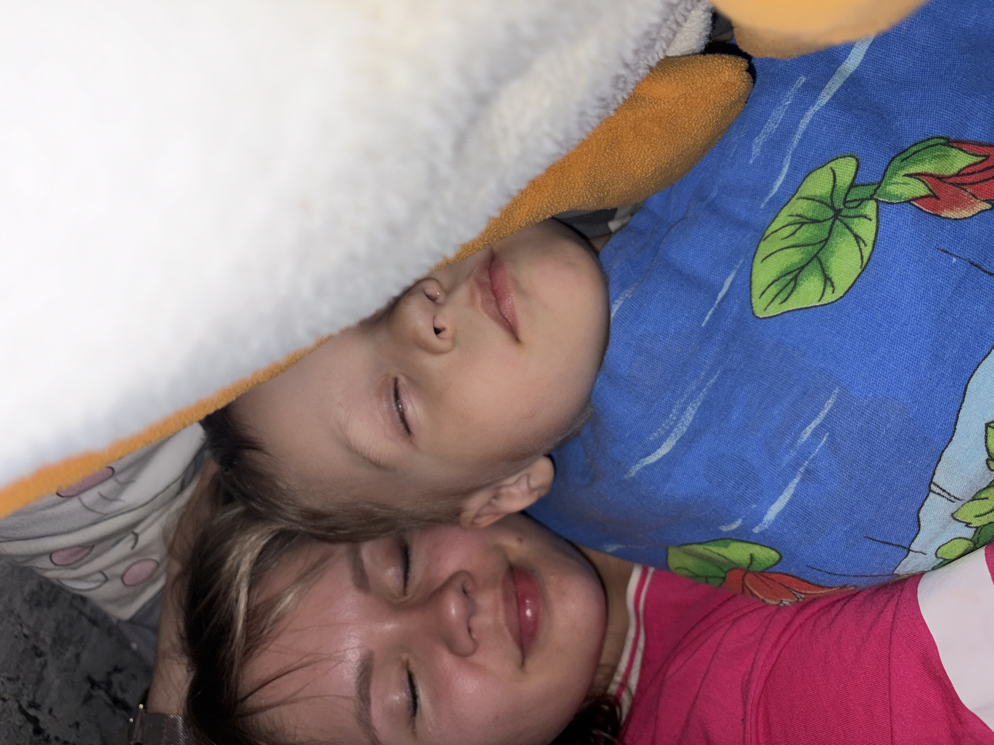 A young woman and a child are lying close together, appearing to be asleep. The child is positioned above the woman, partially covered by a blue pillow with floral and stripe patterns. Above them, there's a white, fluffy material and an orange object, possibly a plush toy. The woman is wearing a pink shirt with white stripes. The location and details surrounding the city of Chaikovsky, Russia, are not evident in the image.