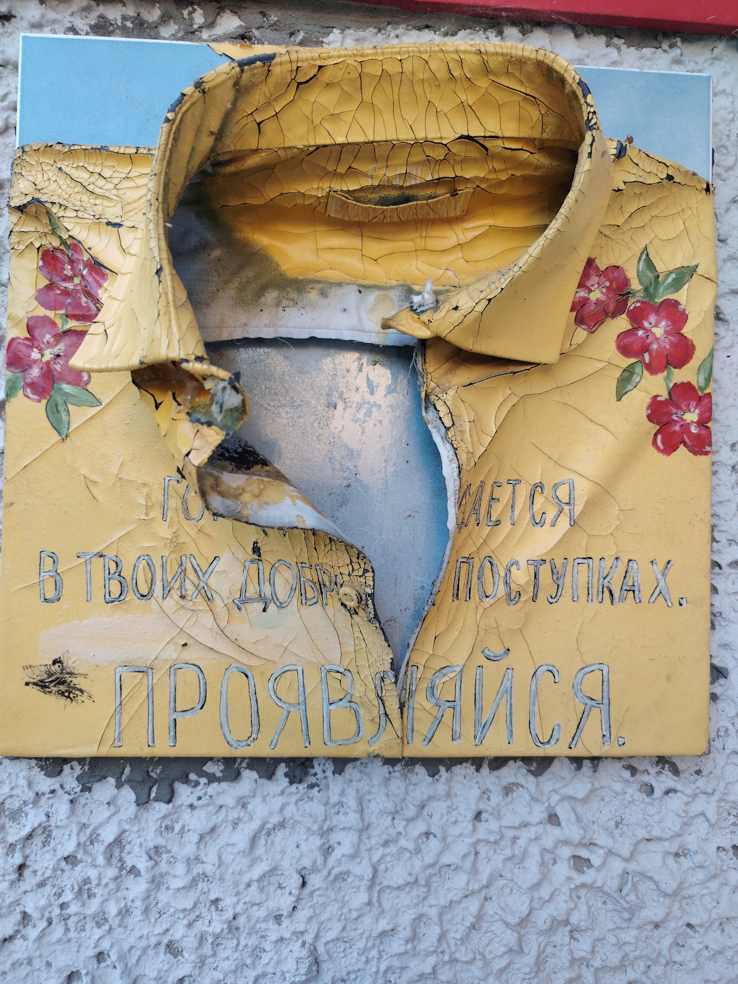 This is a close-up, eye-level, outdoor photograph of a damaged, yellow, shirt-shaped sculpture mounted on a textured white wall. The sculpture is peeling and cracked, with red floral motifs painted on its surface. Cyrillic text is inscribed on the sculpture. The text appears to read "В ТВОИХ ДОБРЫХ ПОСТУПКАХ. ПРОЯВЛЯЙСЯ." This translates to "In your good deeds. Manifest yourself." The sculpture is positioned on a roughly textured, light-colored wall, possibly stucco, which appears to be part of an exterior building surface. The background shows a sliver of a light blue surface, possibly a sky or a painted background element. The overall impression is one of an artistic installation or street art that has weathered over time. The exact location in Konakovo, Russia, is not discernible from the image itself.