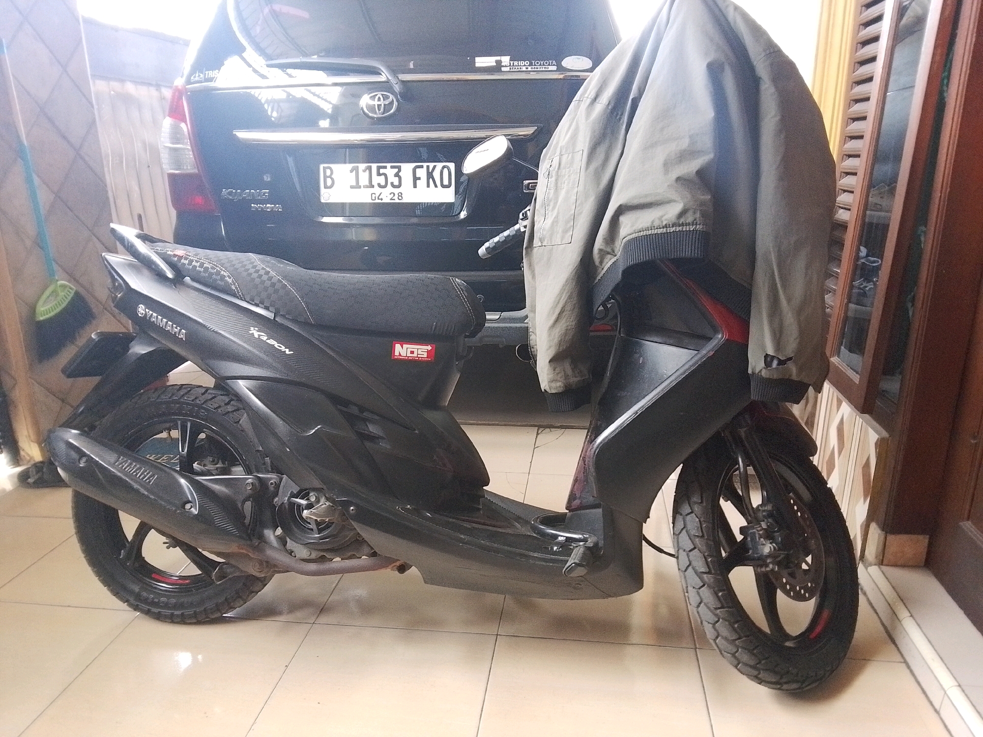 A parked black Yamaha Mio scooter is in the foreground, facing right, with a light green jacket draped over its handlebars and seat. Behind the scooter, a black SUV is parked, facing away from the viewer. The license plate of the SUV reads "B 1153 FKO" and "04-28". The scene appears to be an outdoor or semi-outdoor location, possibly a driveway or garage, with a tiled floor and a wooden door or partition to the right. A broom with a green handle is visible to the left. The overall lighting suggests it is daytime. The presence of the Indonesian license plate indicates the location is in Indonesia, likely within the city of Babelan based on context not present in the image itself.